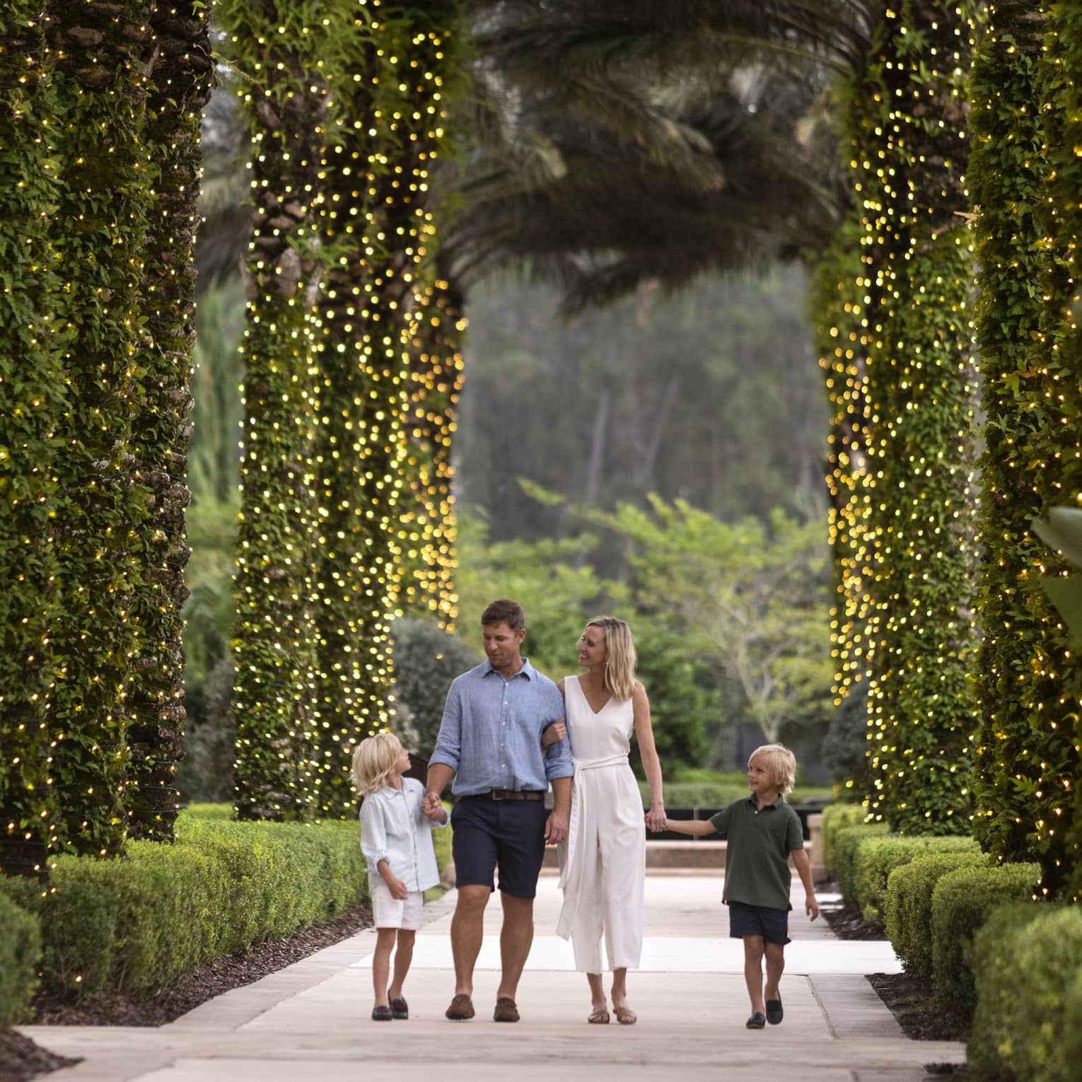 A mother, father and two sons walking along a path of palm trees wrapped in Christmas lights.