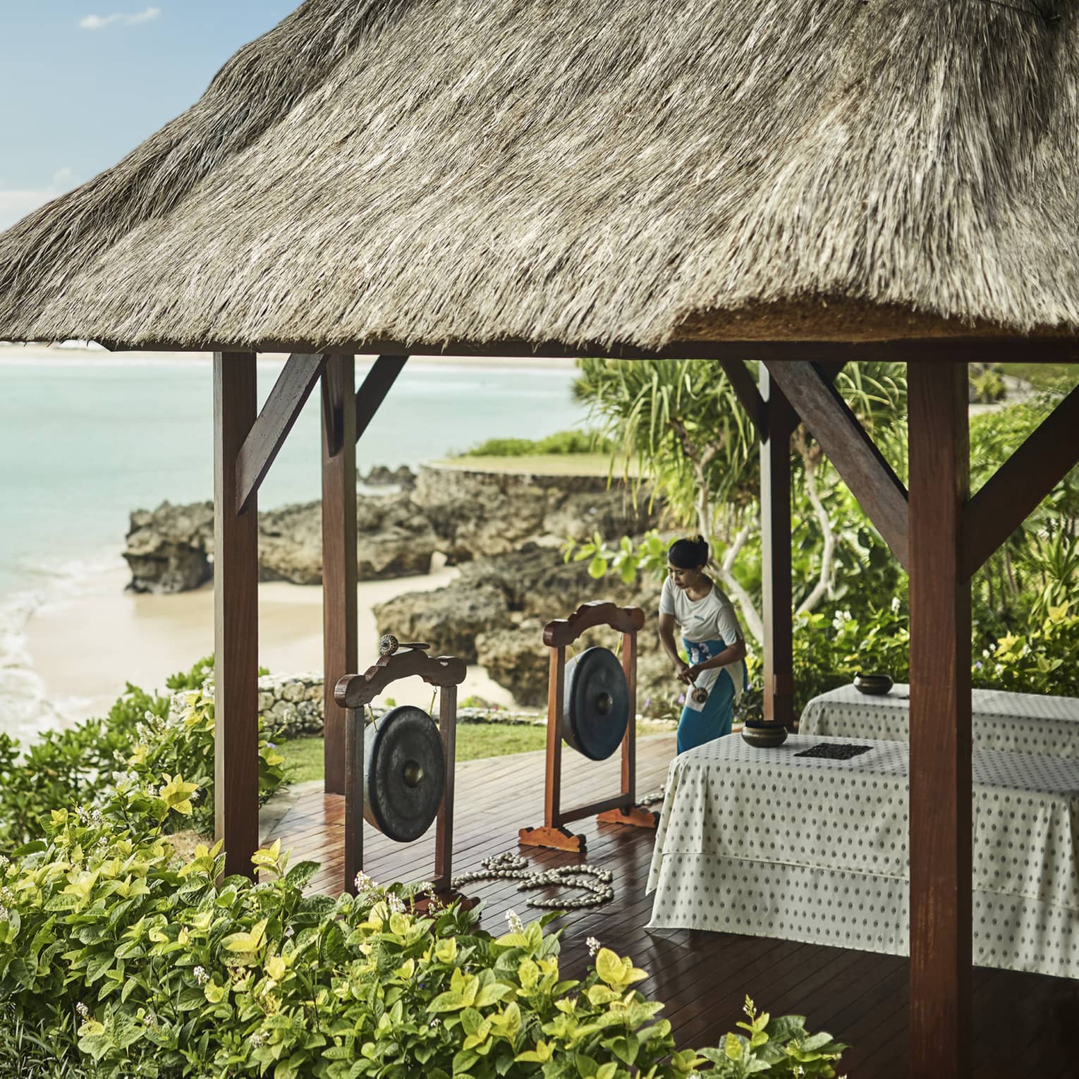 Spa attendant prepares to strike Balinese in front of two massage tables under gazebo overlooking beach, ocean