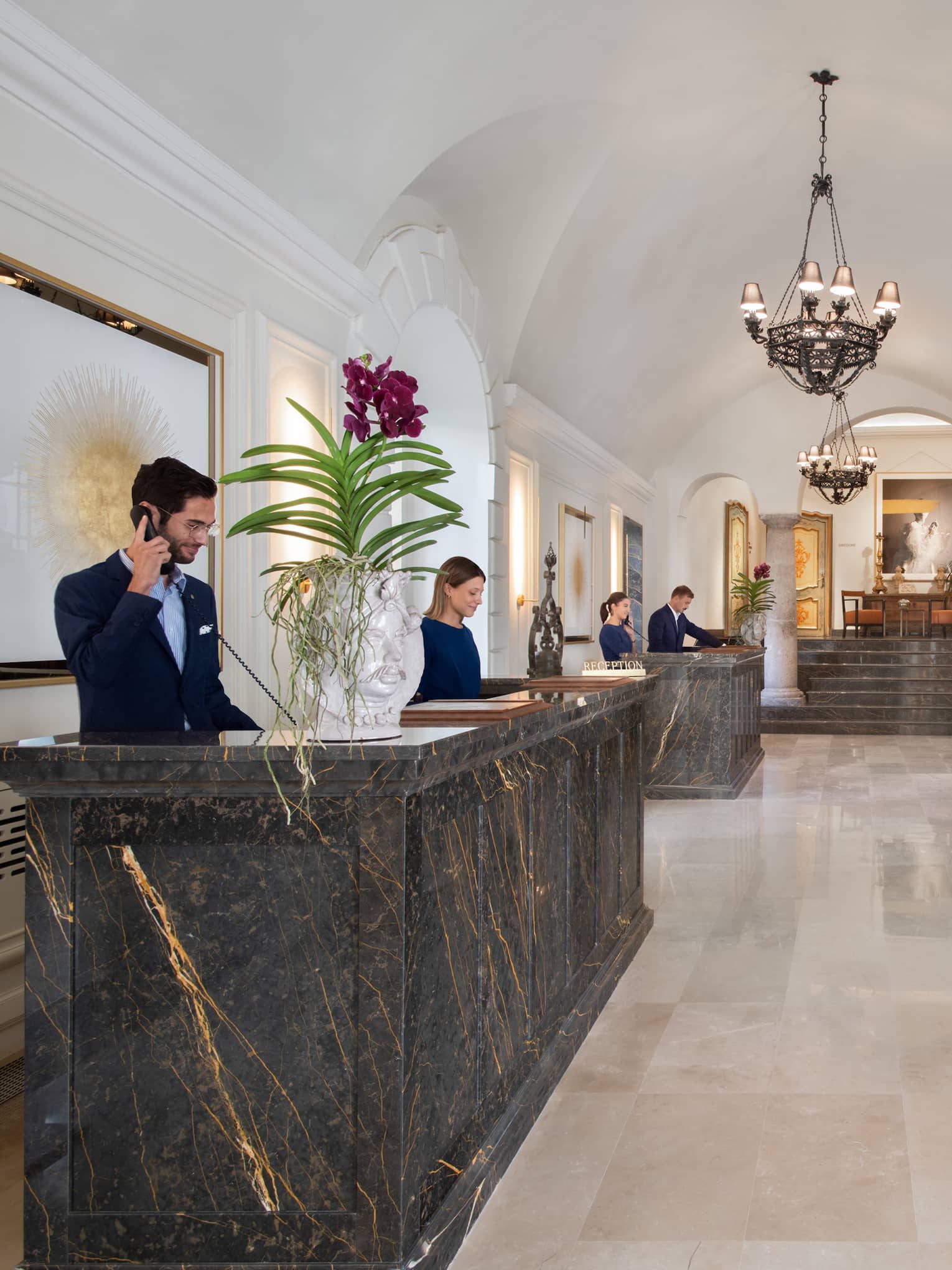 Elegant hotel reception area with people behind marble desks topped with large orchids facing long red velvet and wood bench