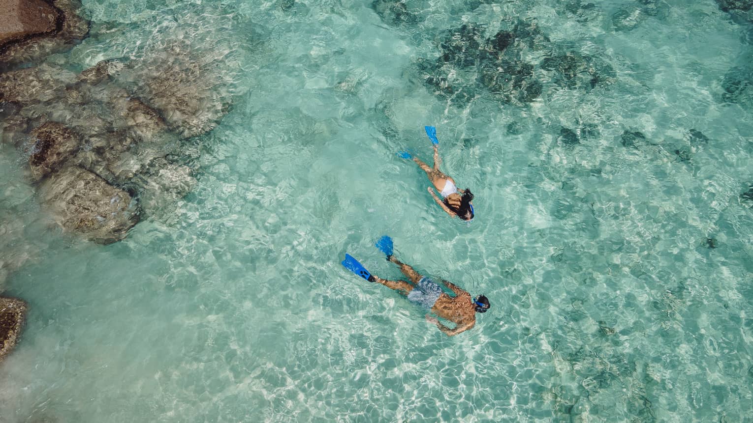 Aerial view of two snorkellers wearing blue flippers in an expanse of shallow, turquoise waters rippling over the reef.