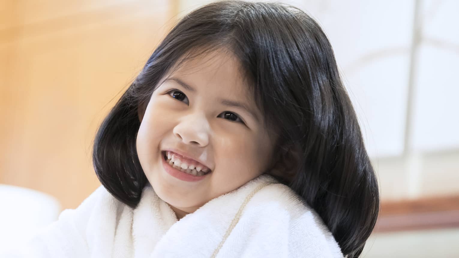 Smiling little girl wearing white bath robe and pink slippers sits on bed
