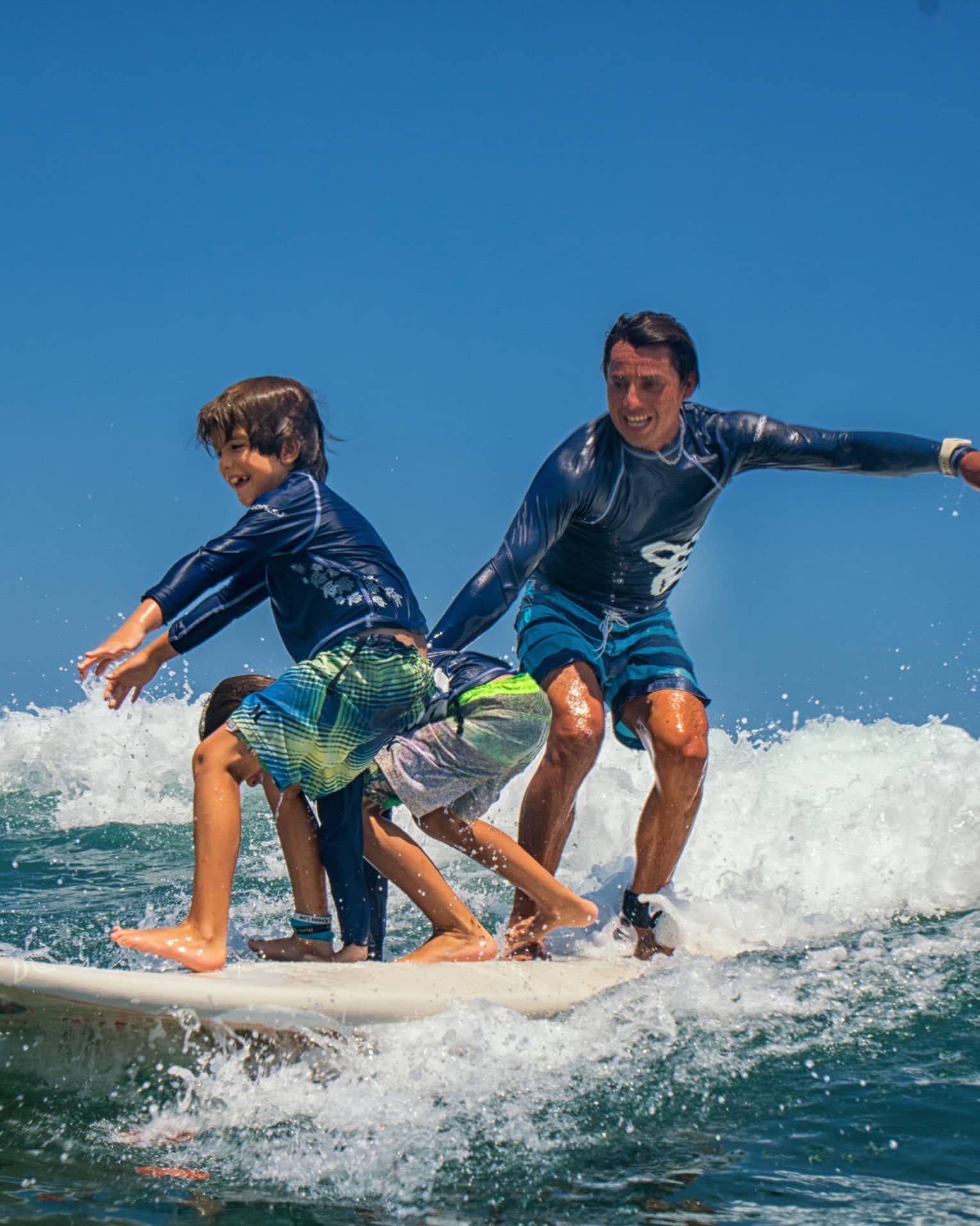 Adult and two children share a surfboard, laughing as they balance on a foamy wave. 