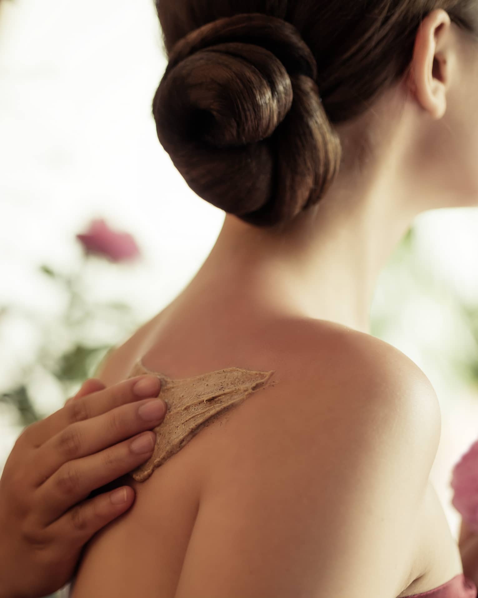 Spa staff rubs thick lotion on woman's bare shoulders as she holds a fresh flower
