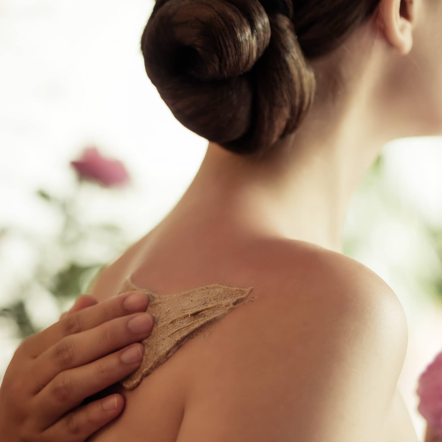 Spa staff rubs thick lotion on woman's bare shoulders as she holds a fresh flower