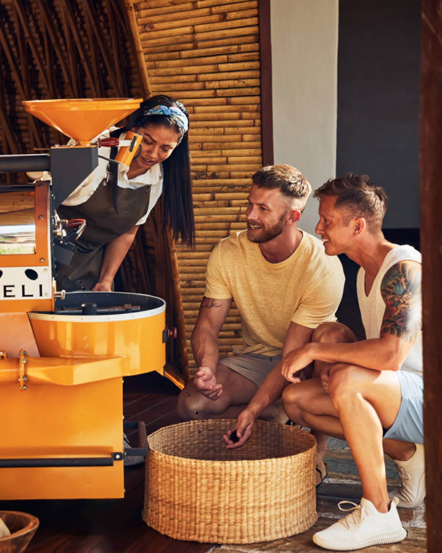 A woman demonstrates coffee roasting to two guests, who observe closely near a yellow coffee roaster in a bamboo-walled setting.