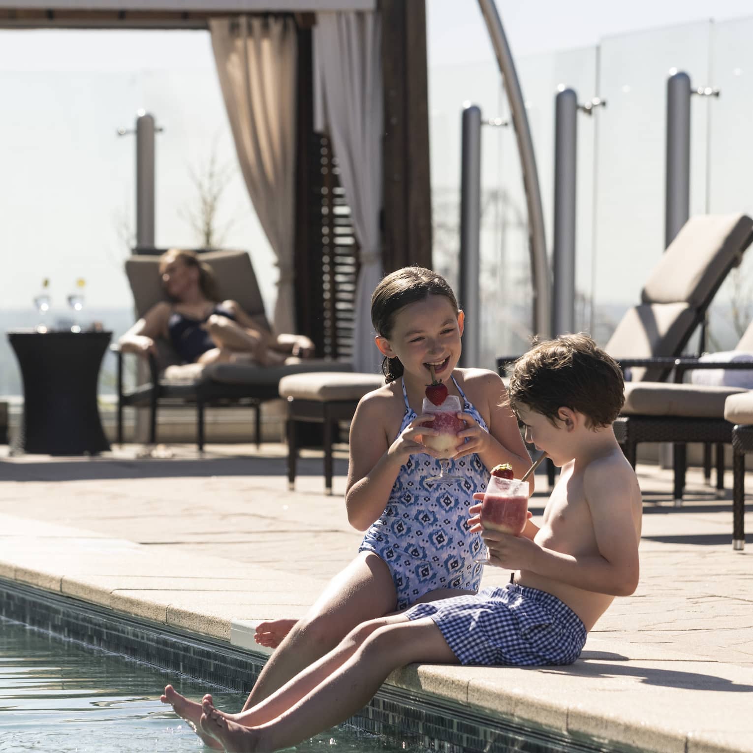 Two children sitting by the edge of a pool, enjoying drinks, while adults relax on lounge chairs under shaded cabanas in the background