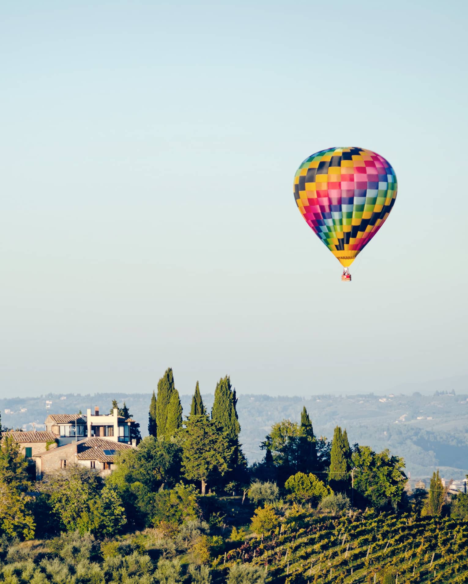 Colourful hot air balloon floating above a landscape with trees and houses, set against a clear blue sky.		