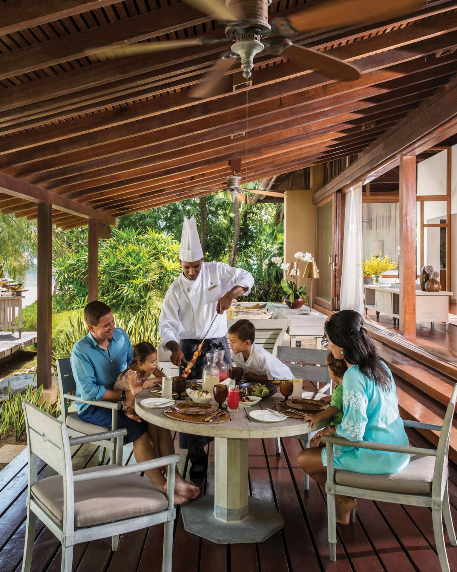 Chef in uniform presents meat skewer to family around table on Beach Villa patio