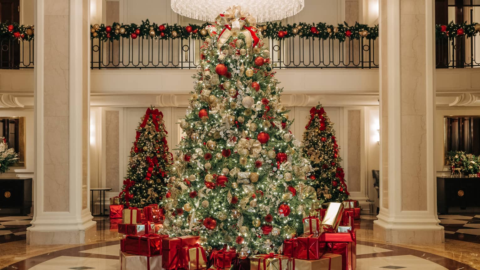 Tall Christmas tree decorated in gold, silver and red with presents all around sits in the centre of a hotel lobby, with two smaller trees in the the background and garlands draped over a railing