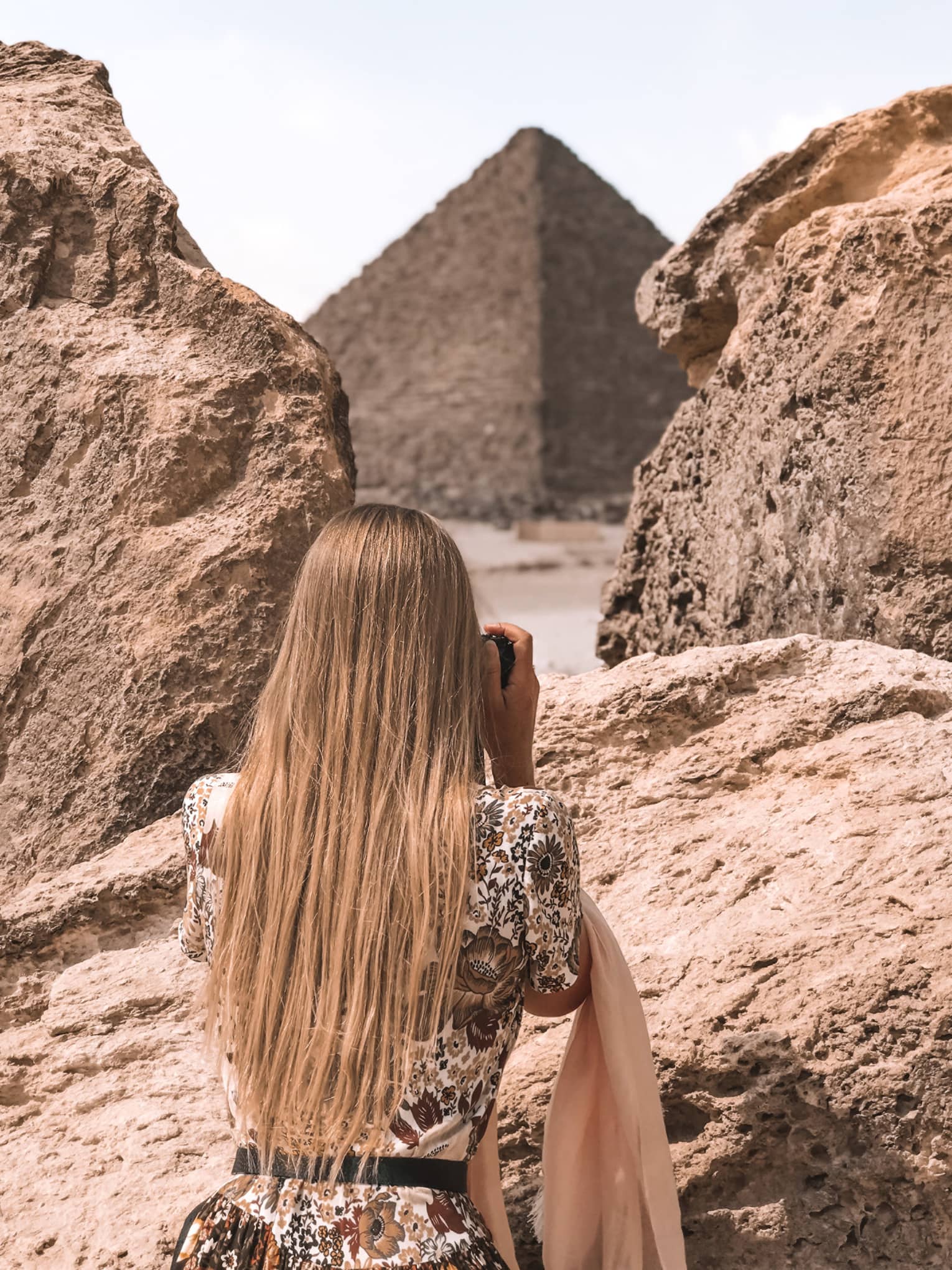 A tourist in a floral dress stands between two large rocks and aims her camera at an enormous pyramid beyond. 
