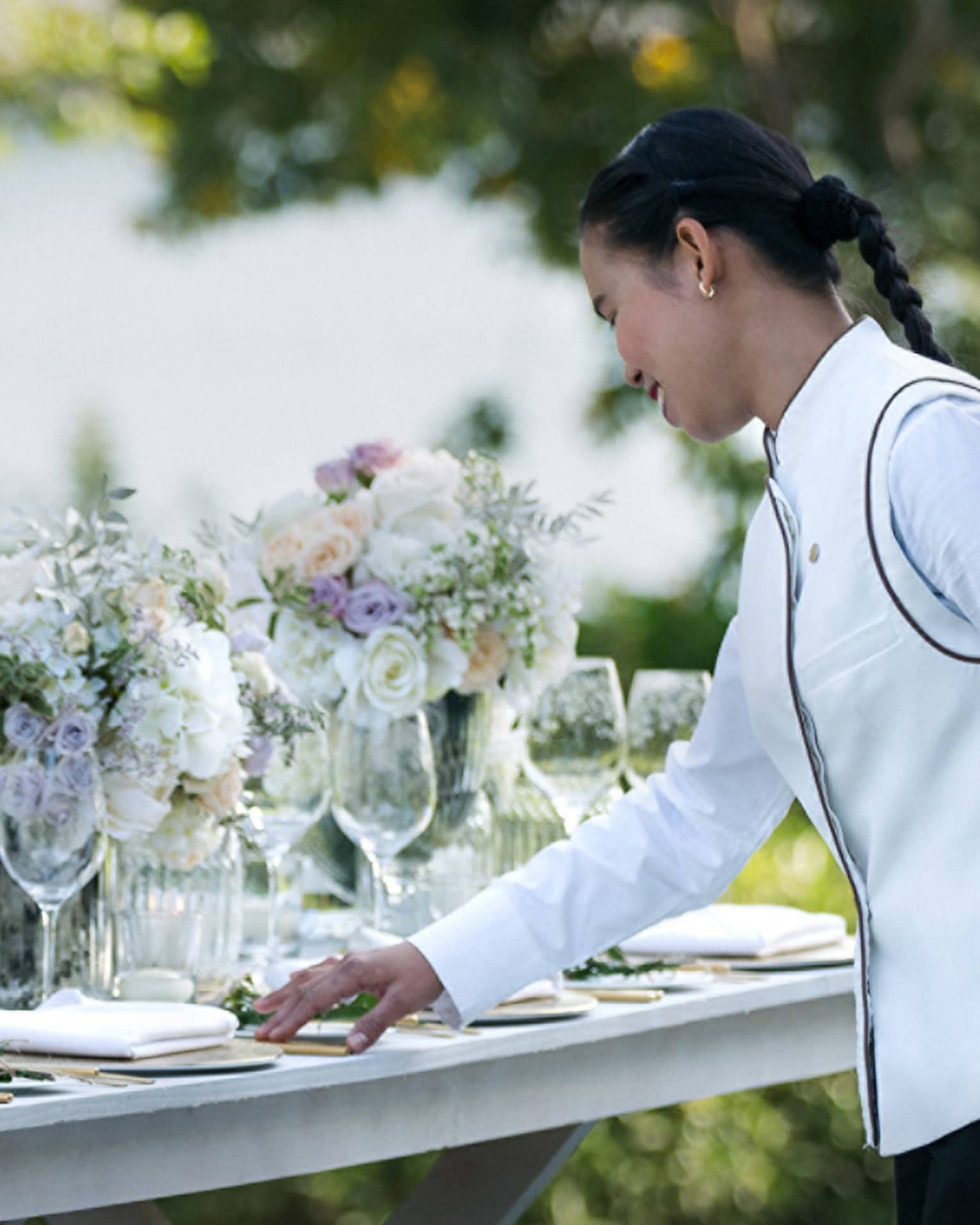 A four seasons staff does the final touches on an outdoor, long banquet table decorated with large white floral arrangements 