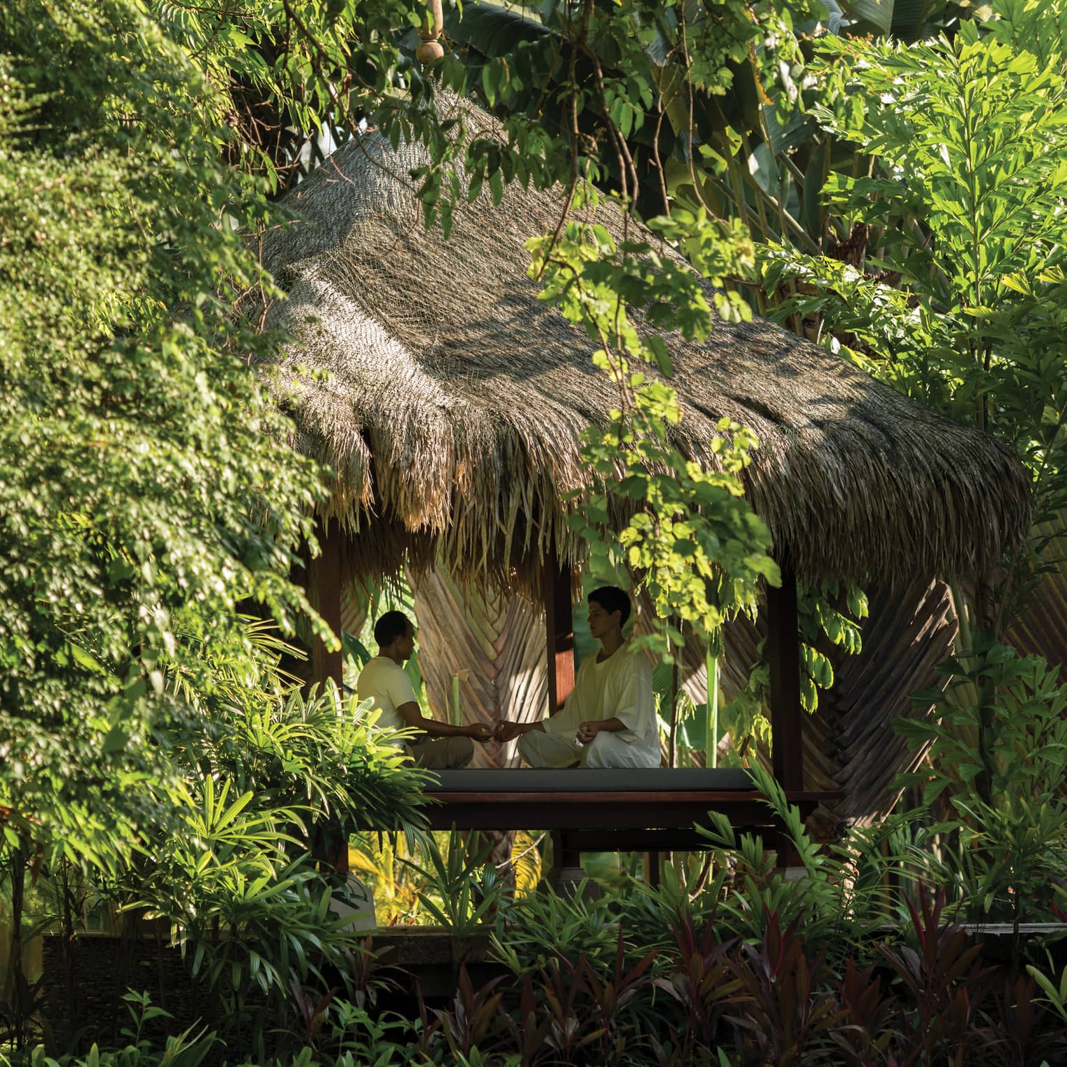 Man and woman sit cross-legged, meditate on wood platform under small hut in forest