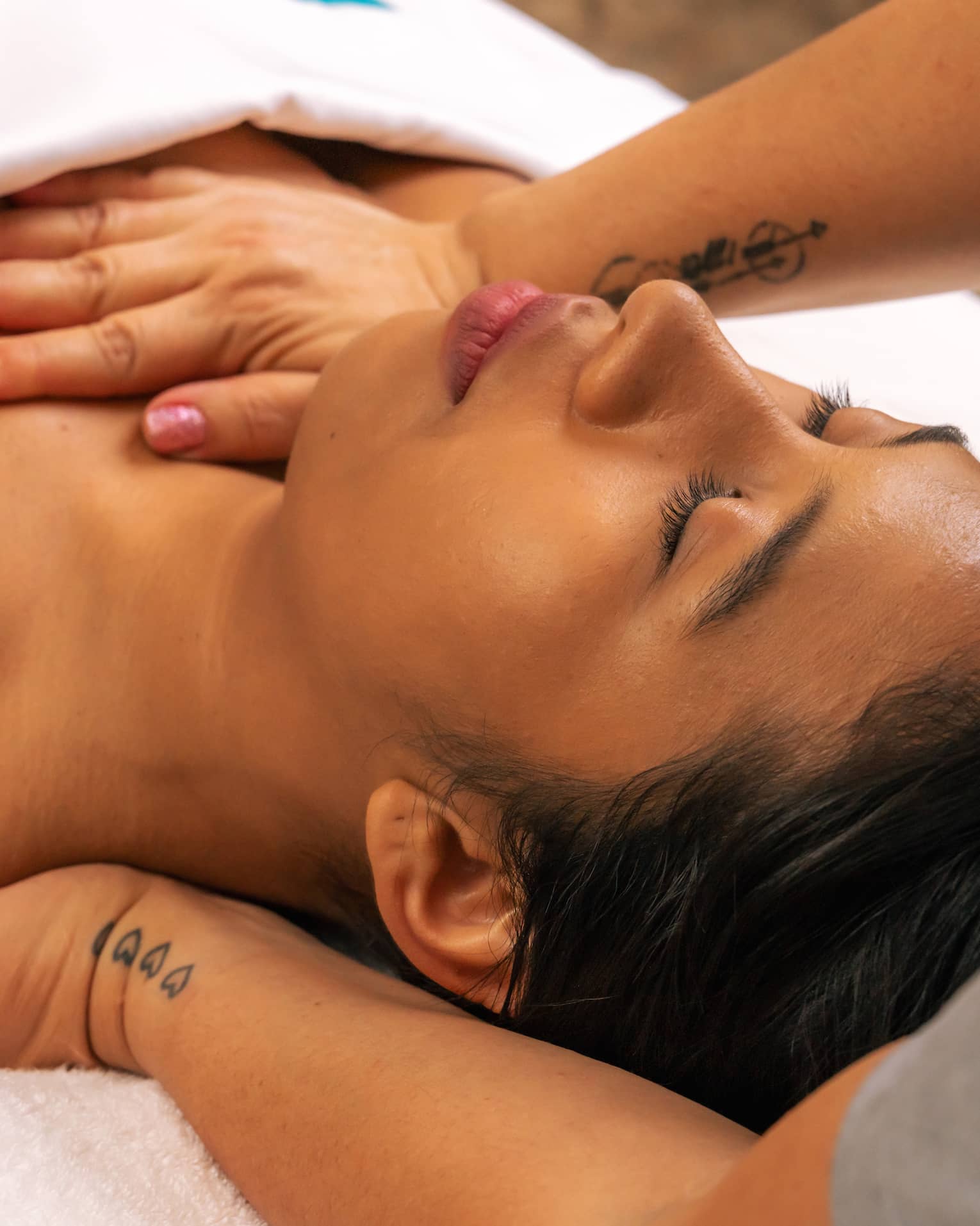 Person lies on their back beneath a white blanket with their eyes closed as a pair of hands massages the back of their shoulder and applies pressure on sternum