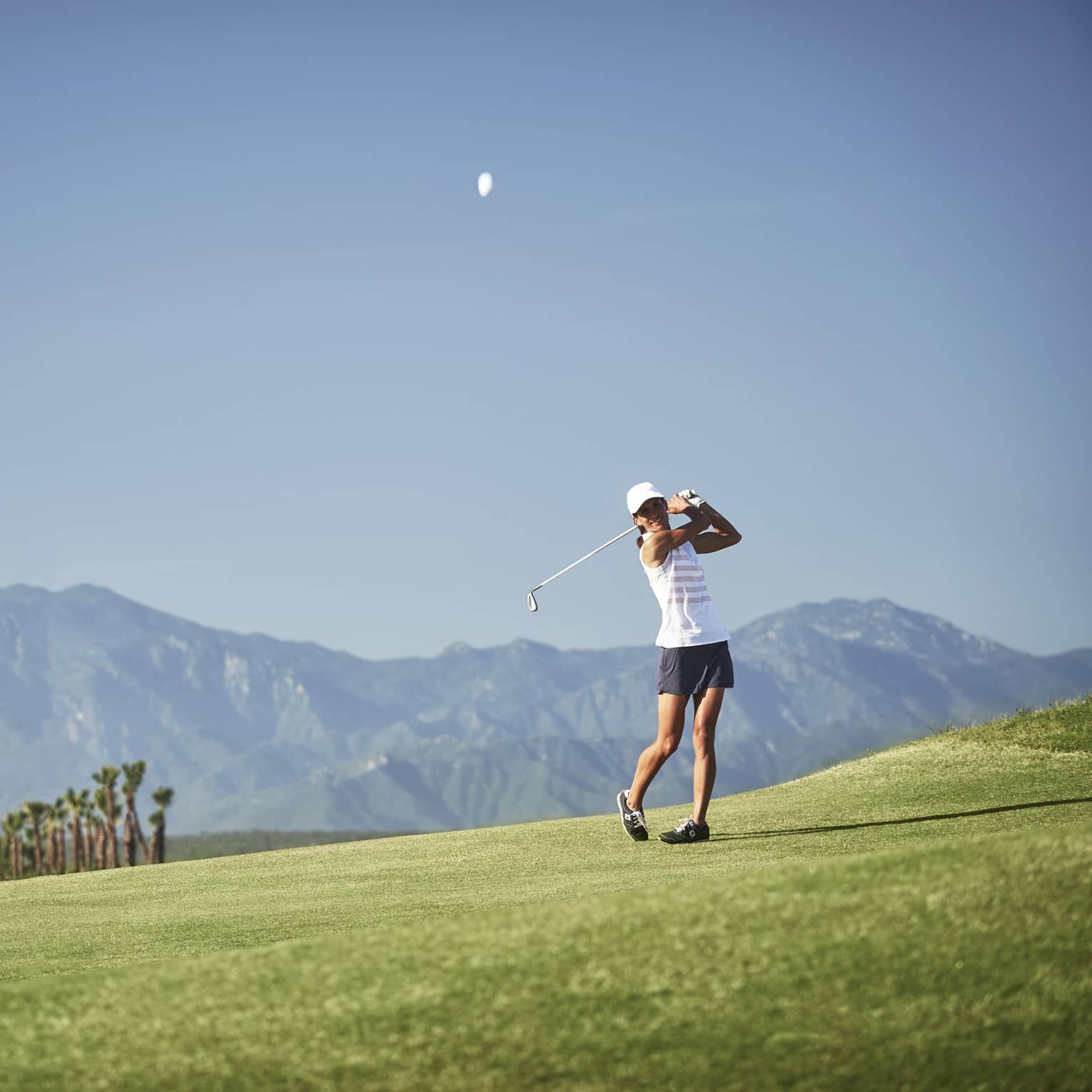 On a smooth fairway against a striking panorama of mountains, a golfer ends their swing with a confident follow-through.