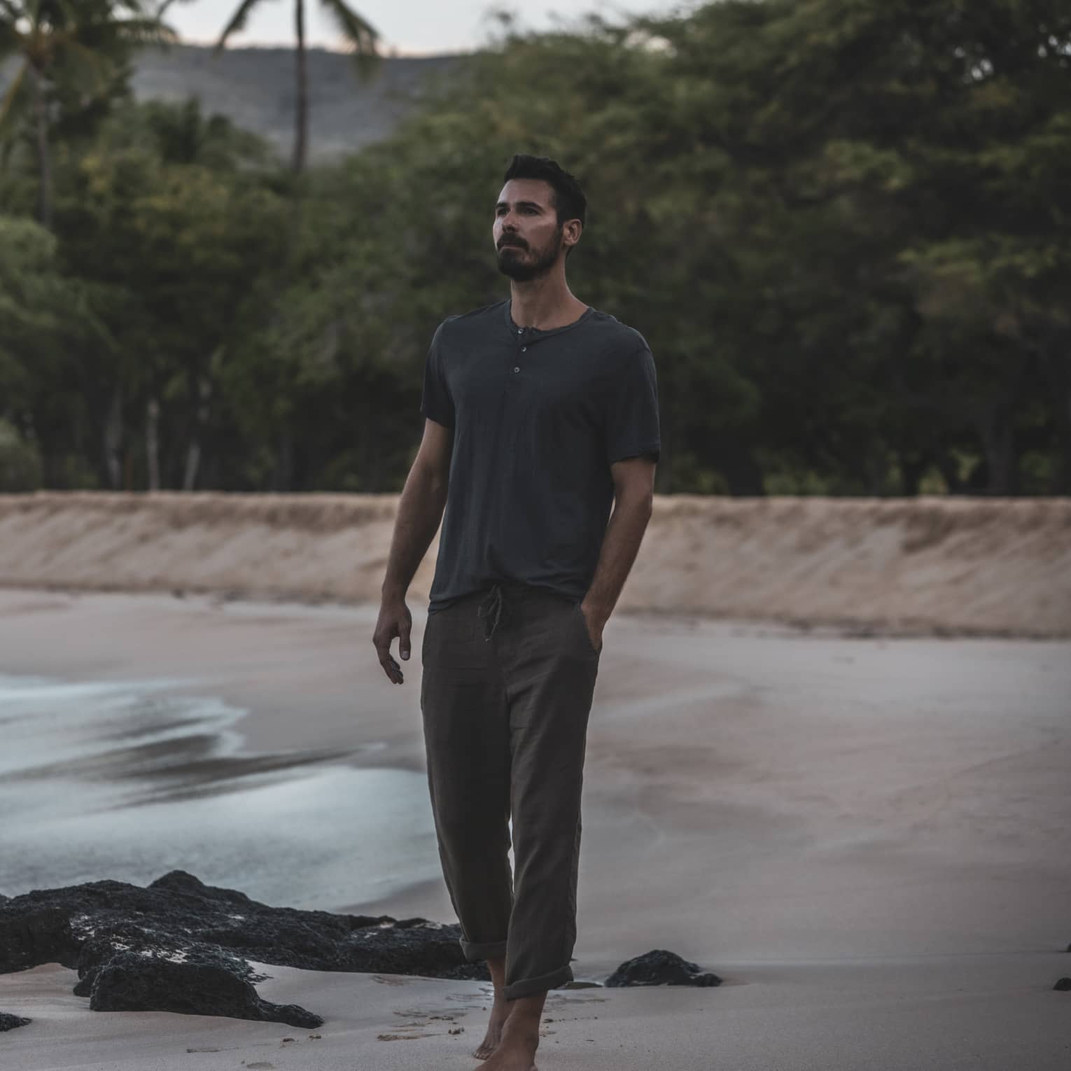 A man walks on a beach in front of a lush tropical forest