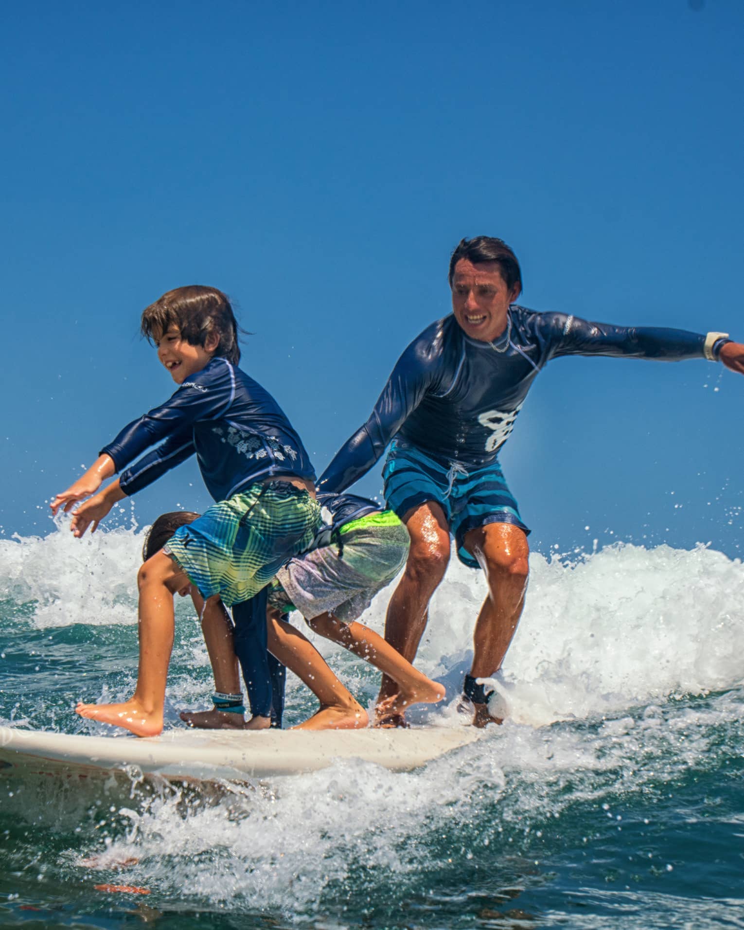 Adult and two children share a surfboard, laughing as they balance on a foamy wave. 