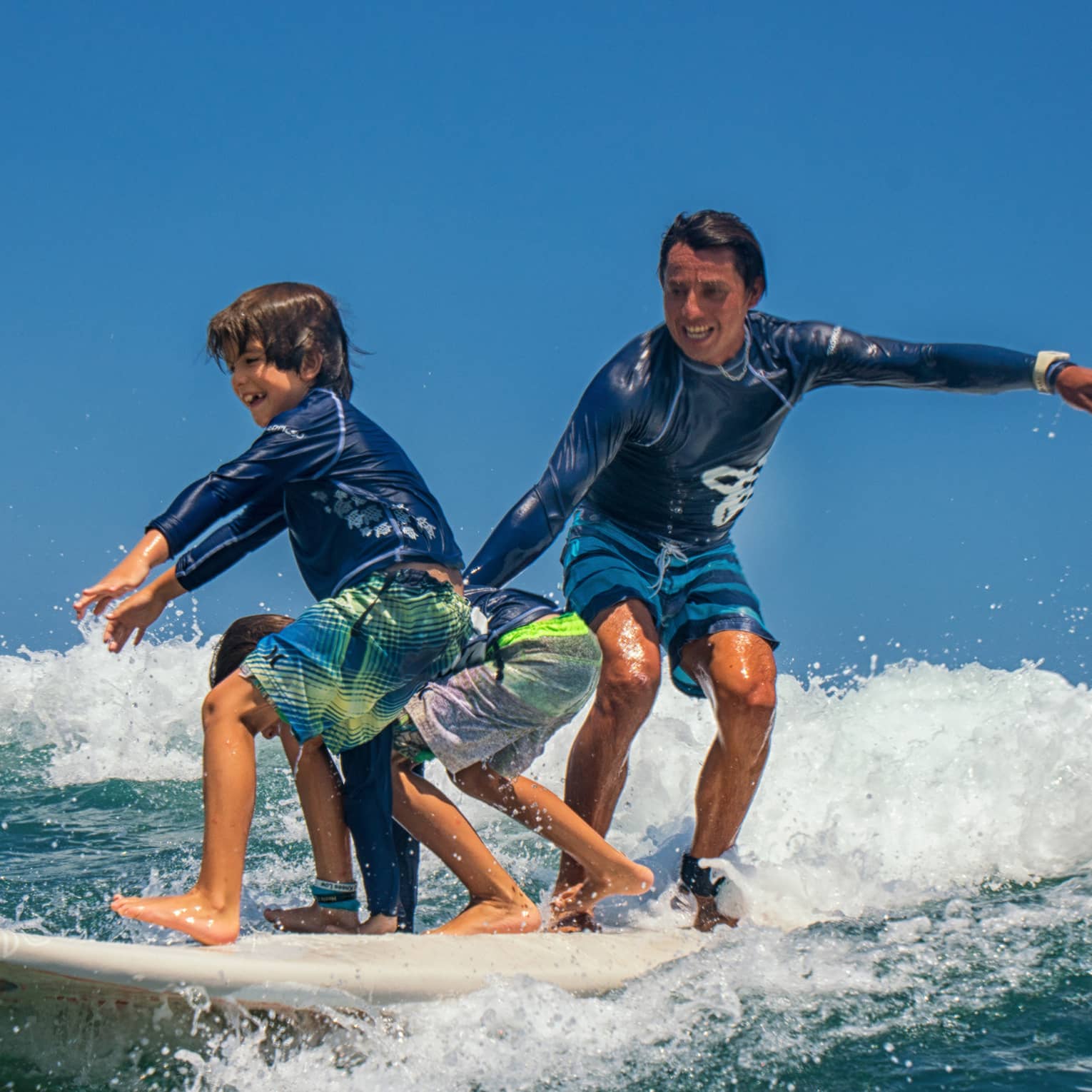Adult and two children share a surfboard, laughing as they balance on a foamy wave. 