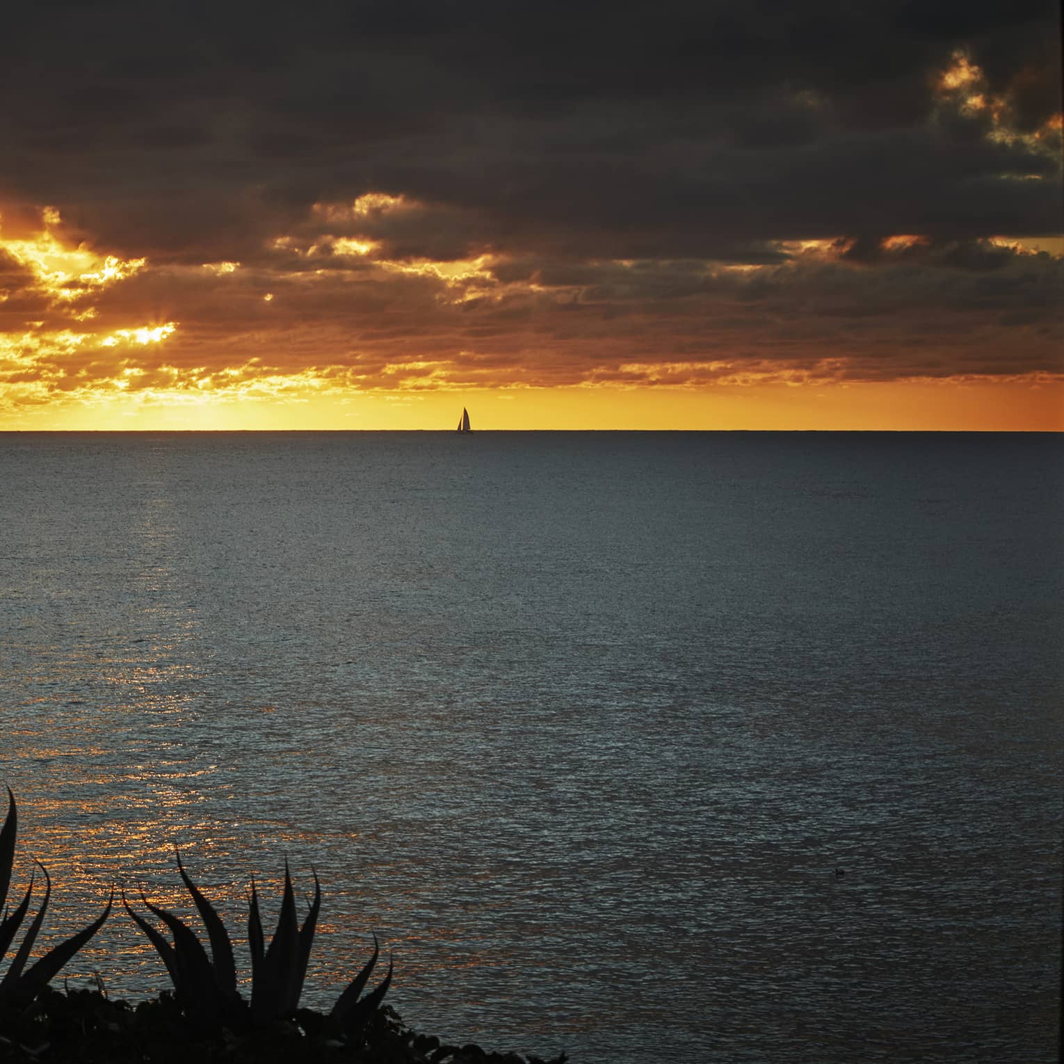 Bright orange sunset over the ocean with dramatic clouds and a sailboat in the distance