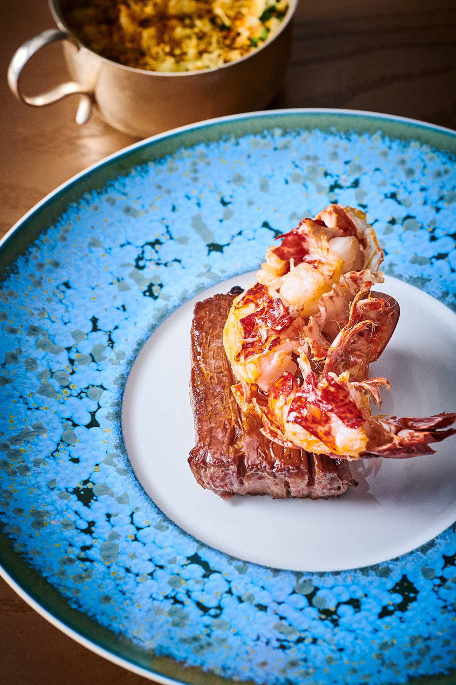Half-lobster in shell atop a beef fillet on white plate with speckled blue rim