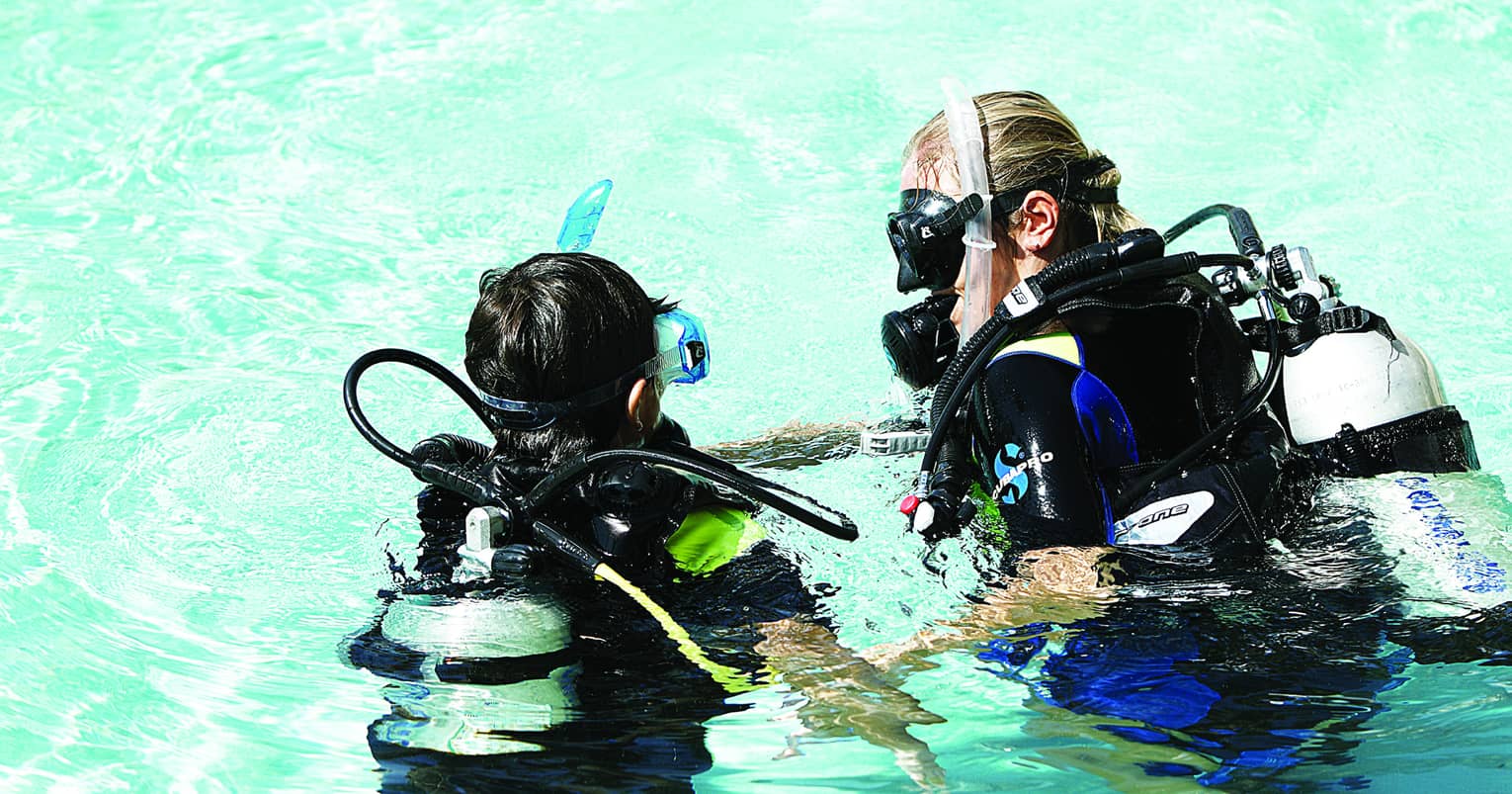 An instructor guiding a child, both in wetsuits and full scuba gear, to slowly submerge into glistening turquoise water.