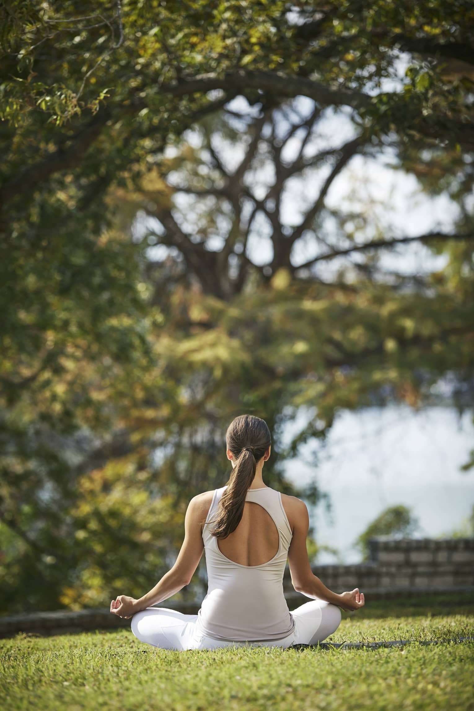 Rear view of a guest in white athletic wear meditating cross-legged with forearms rested on knees on a sunny patch of grass.