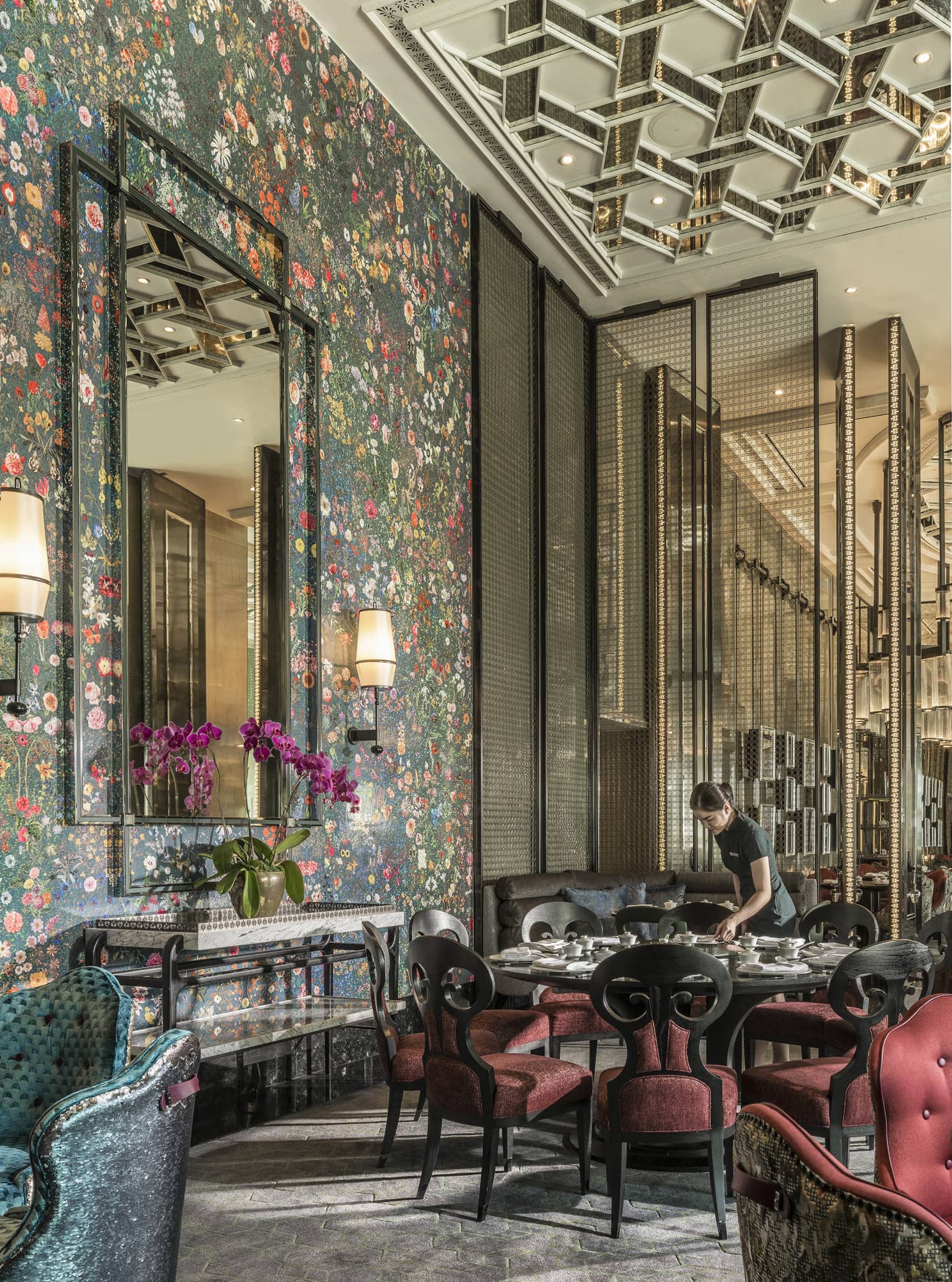 Hotel staff sets table with elegant red velvet chairs under tall mirror, floral wallpaper 