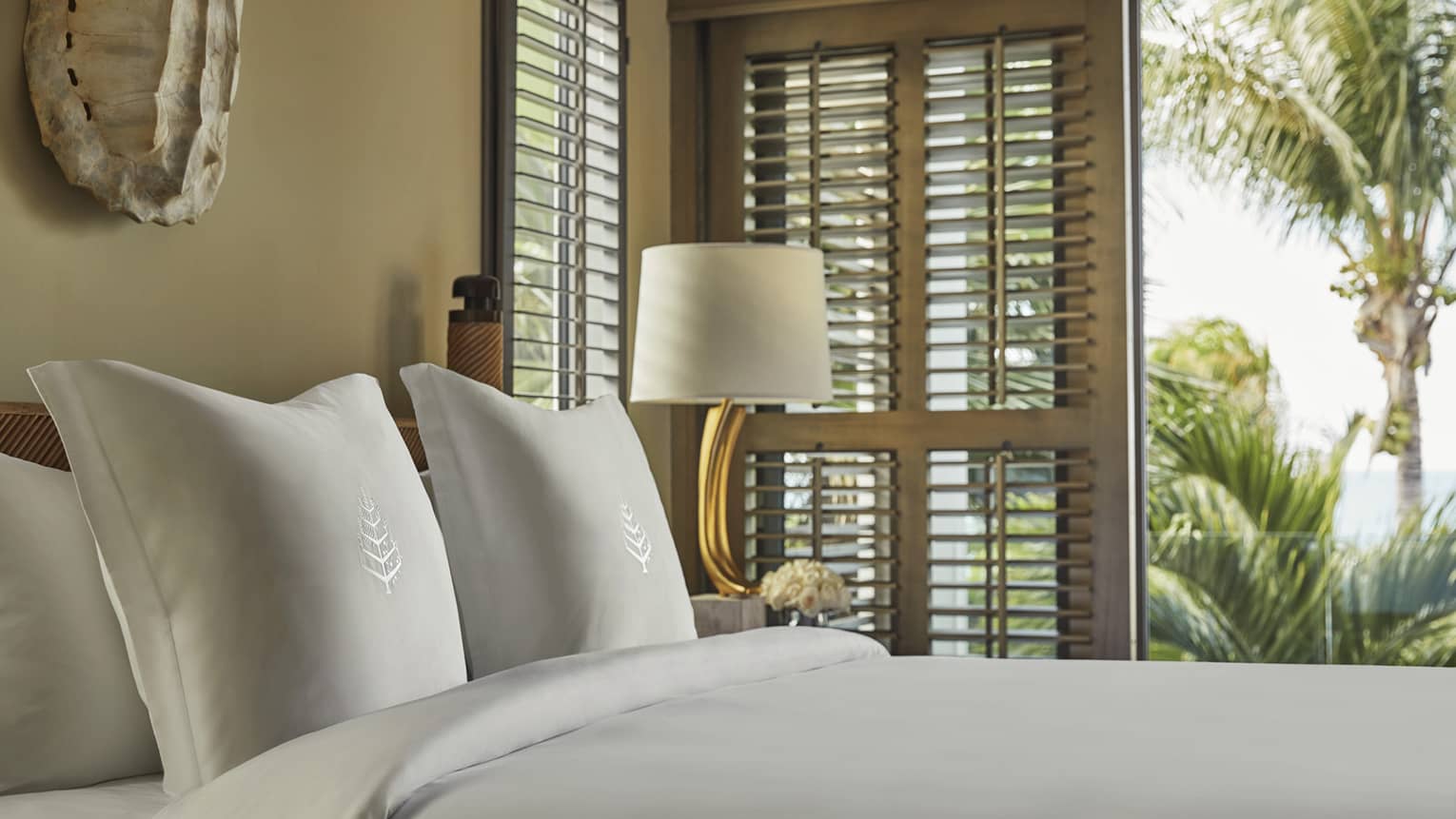 Close-up of a neatly made bed with embroidered white pillows, next to a lamp and window shutters revealing tropical palm trees outside