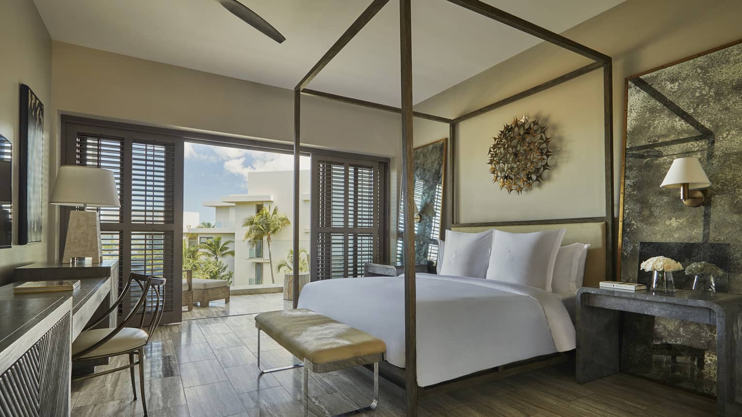 Elegant bedroom with a four-poster bed, desk, neutral tones and balcony doors offering a view of palm trees and blue skies