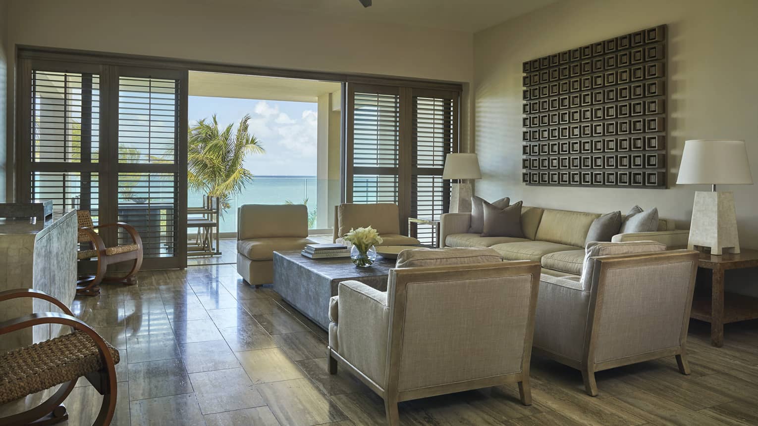 Elegant living area of a resort suite with neutral-toned furniture, modern decor and doors opening to a balcony with ocean and palm tree views