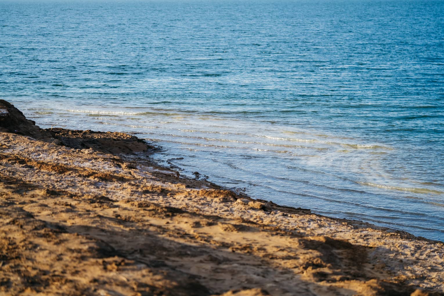 Gentle waves in sparkling azure water undulate toward an empty dark-brown, rough-sand beach.