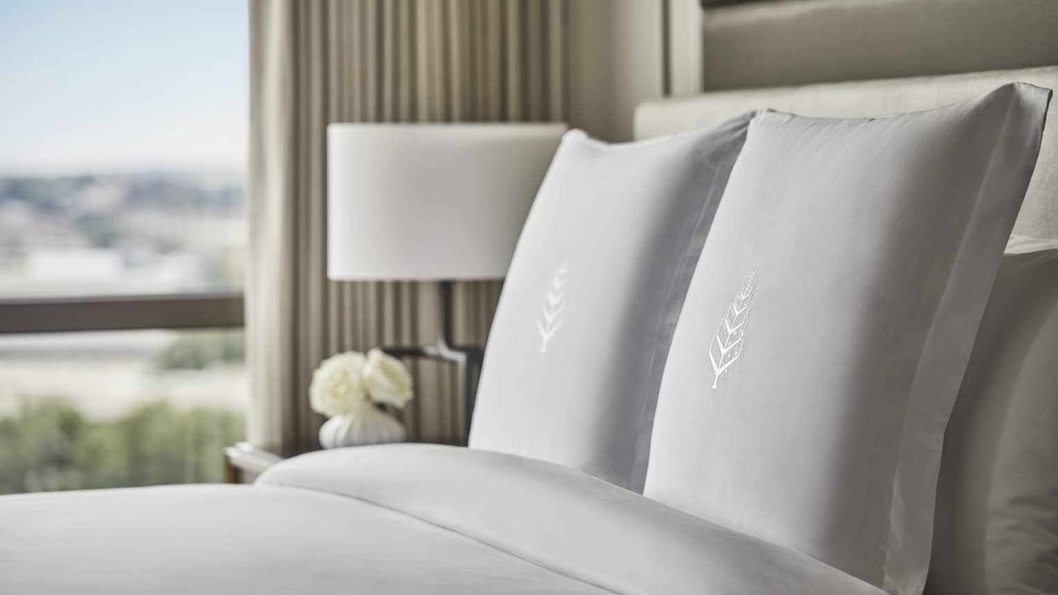 Close-up of hotel bed, pillows with embroidered Four Seasons logo, nightstand with white roses