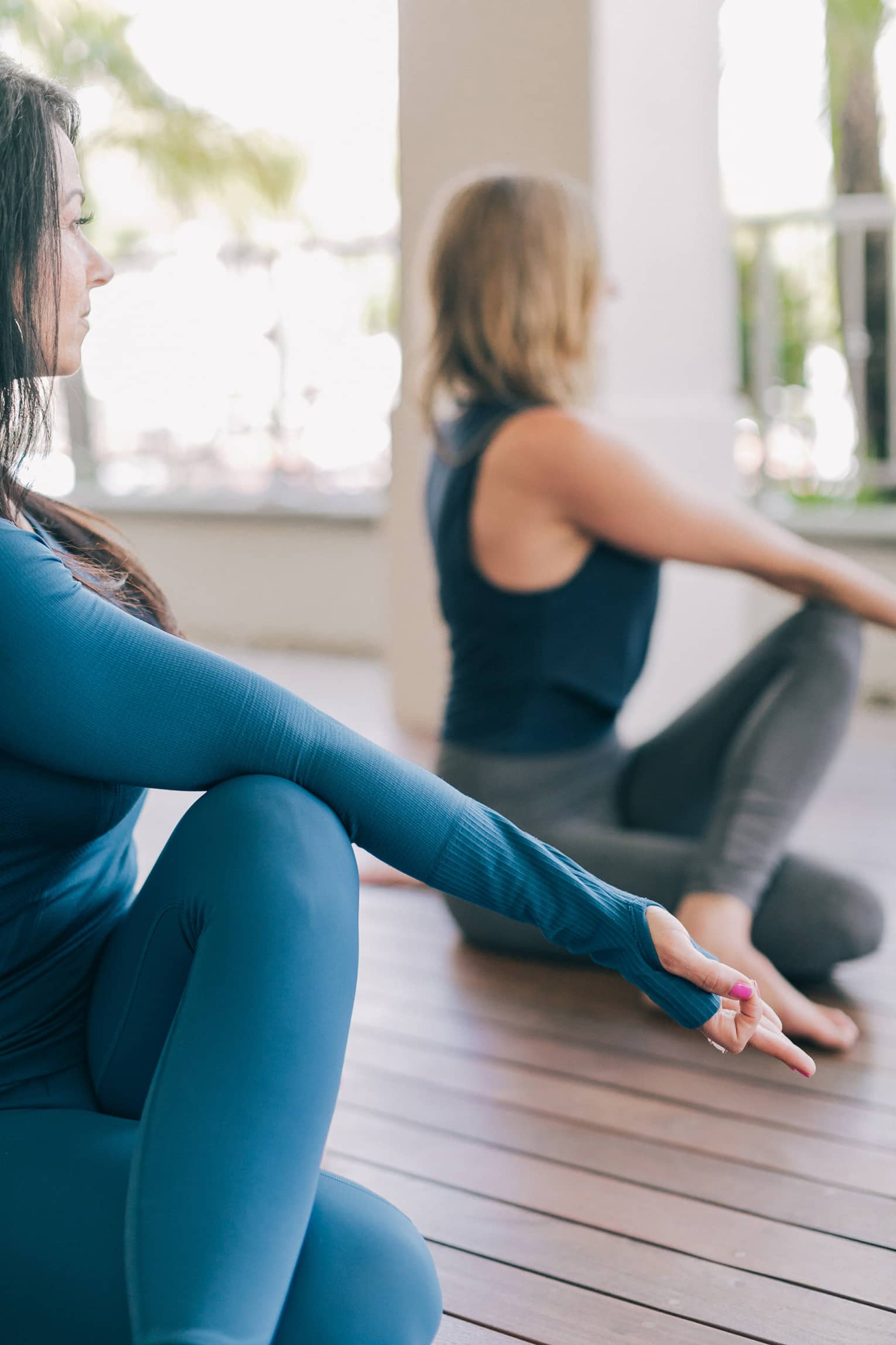 Two women sit on wood plank floors, do yoga stretch