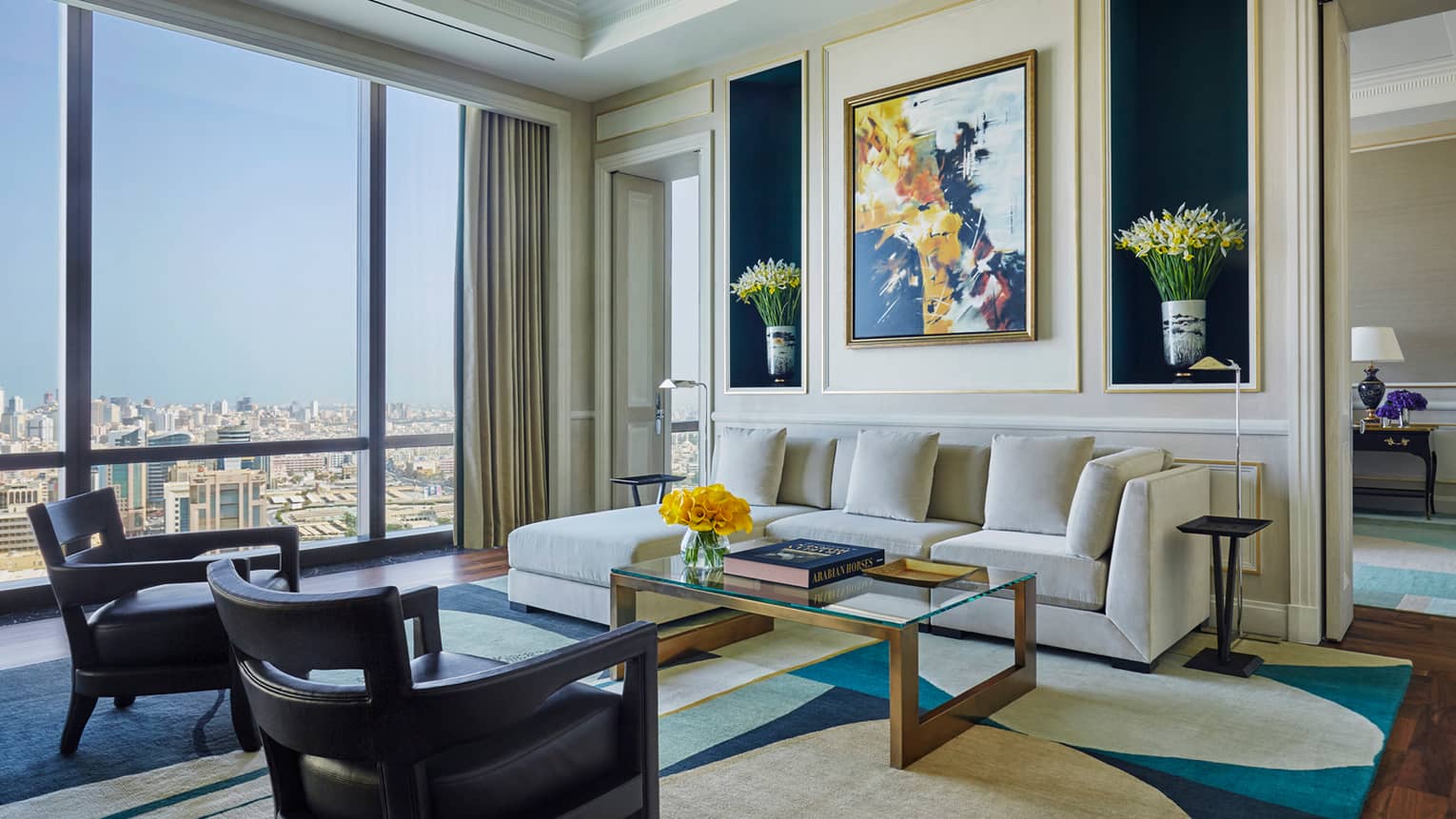 Hotel room living area with L-shaped white sofa, two small armchairs, coffee table with books, colourful painting, flowers