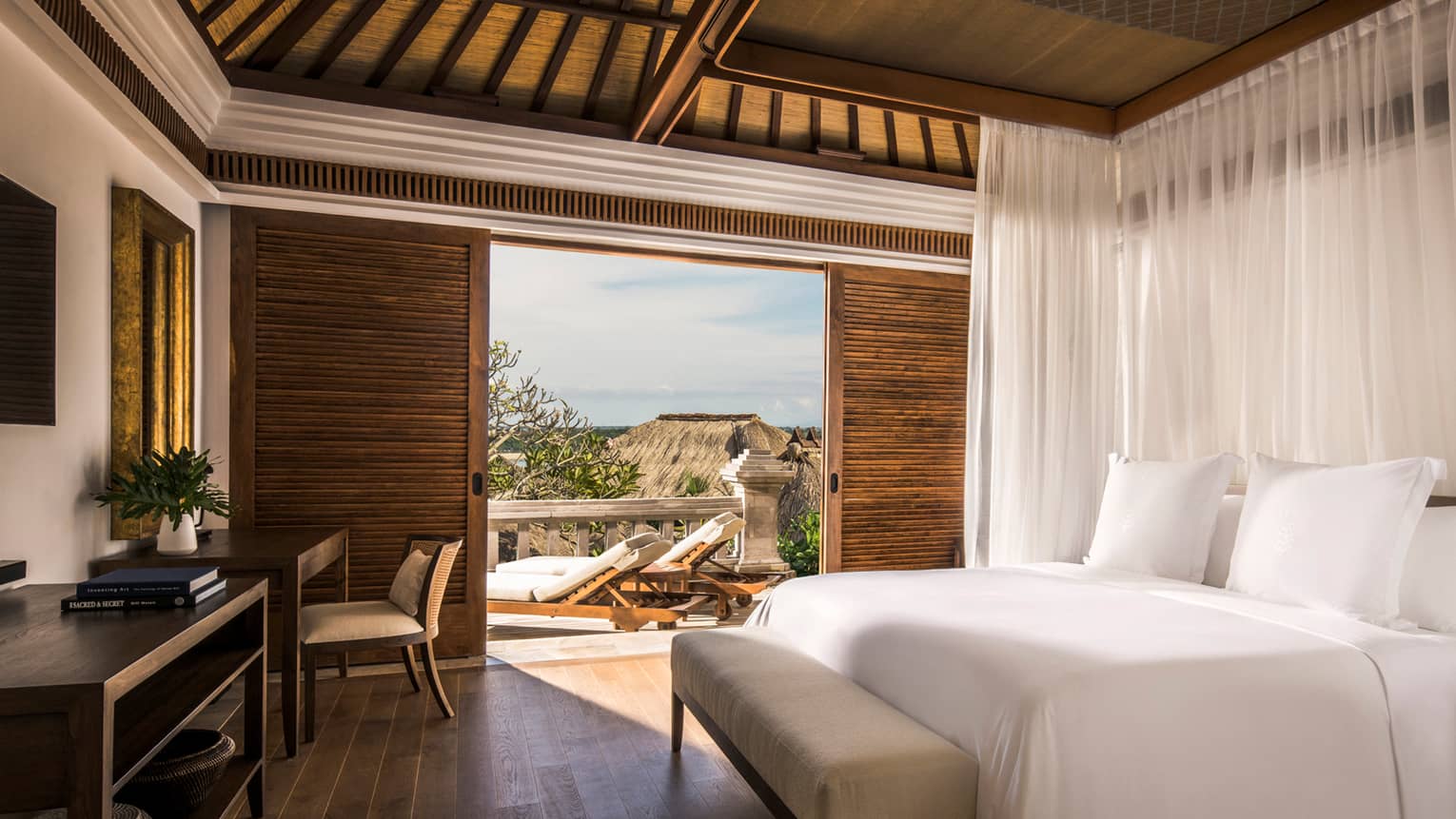 Garden villa bedroom bed, small bench, wood desk beside sliding wood shutters to sunny patio with garden view
