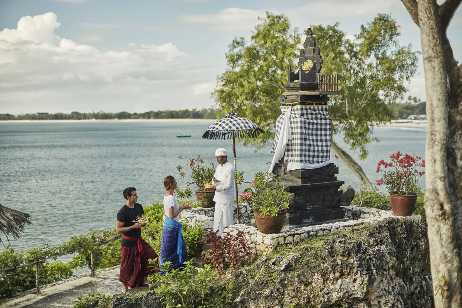 High Priest Aji Ngurah stands by shrine with black-and-white checkered umbrella, cloth, as man and woman make offering