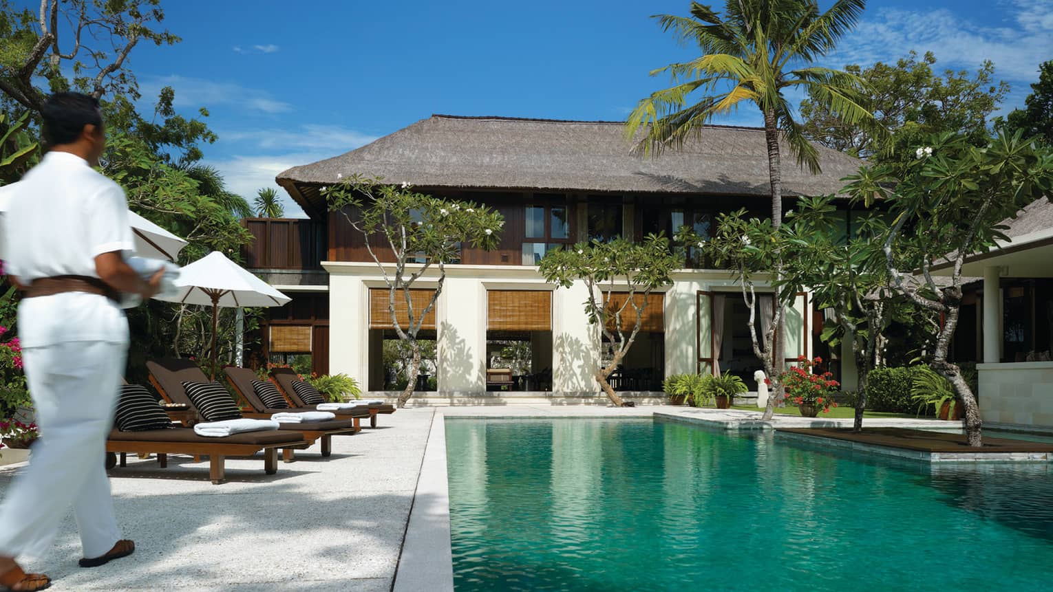 Hotel staff wearing white uniform walks past outdoor pool to villa