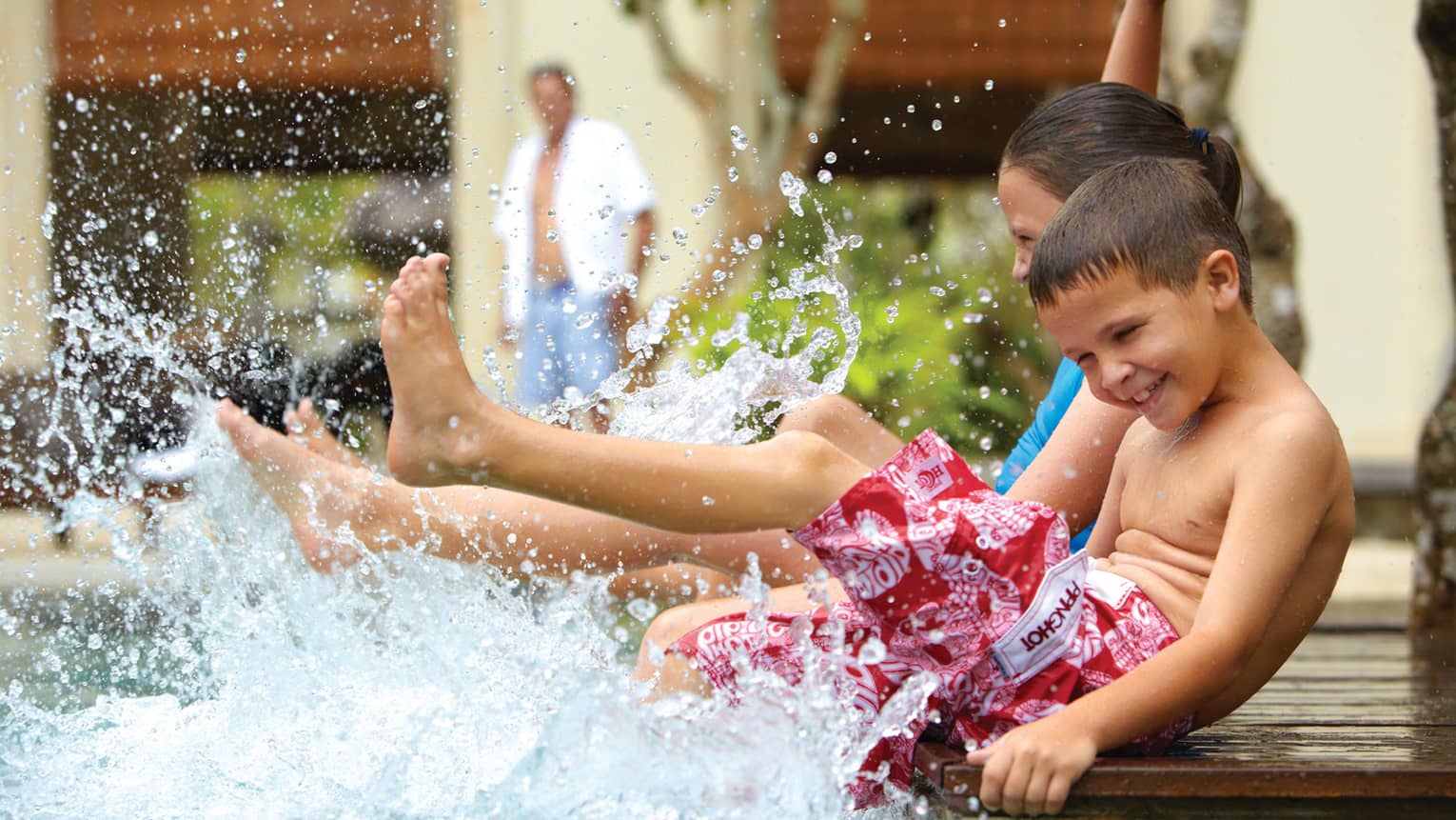 Young children in swimsuits sit on edge of swimming pool, splash water with legs