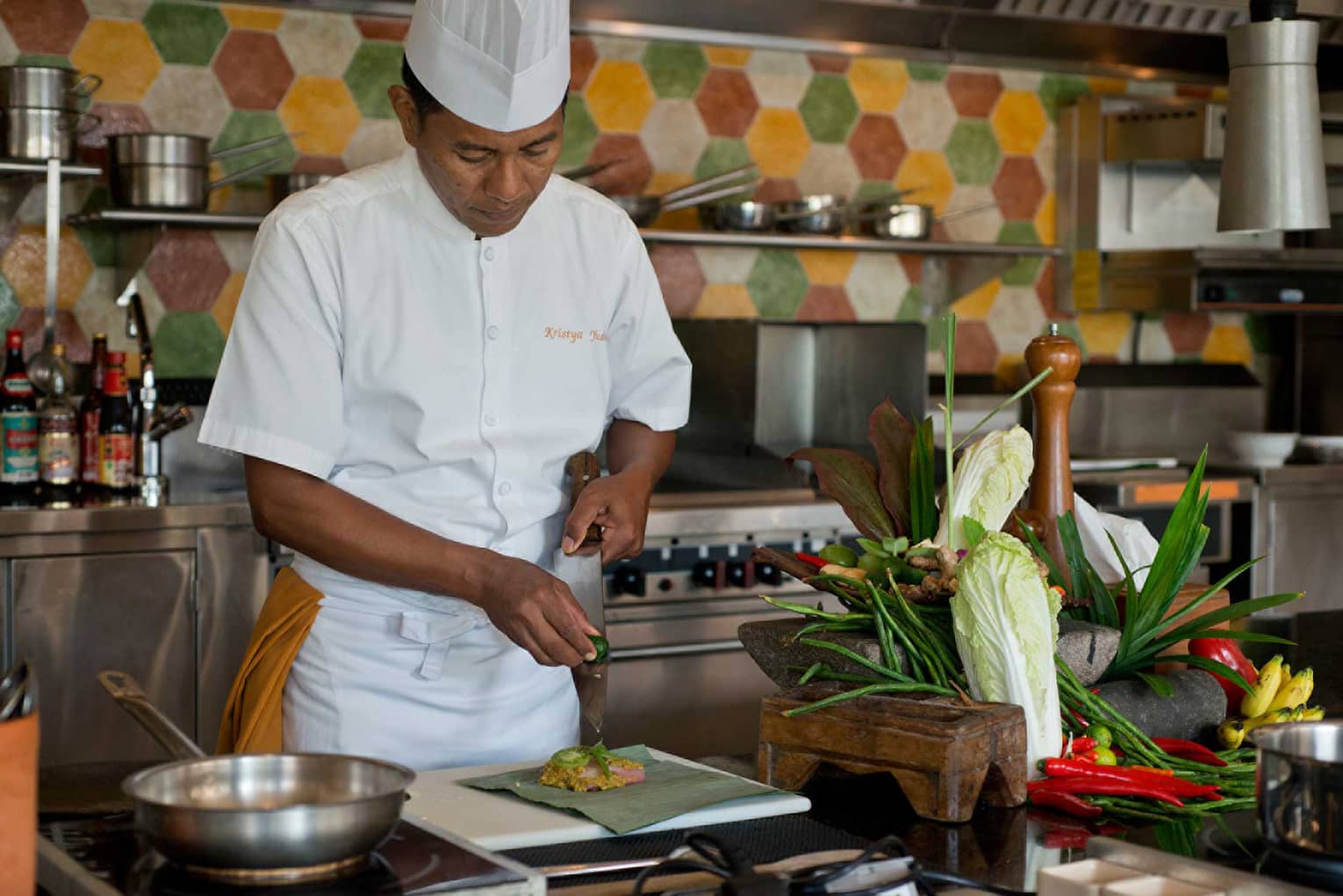 Chef in white uniform squeezes lime on large kitchen knife over dish, next to heads of fresh lettuce and whole peppers