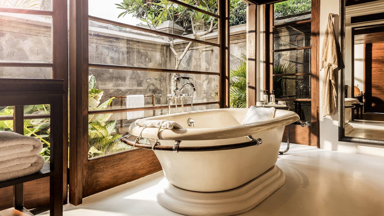 Free-standing bath tub with folded white towels, bathrobe hanging above, large window to garden