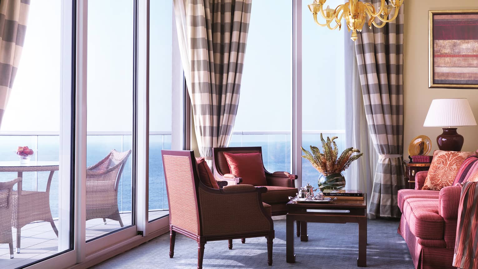 Rose-coloured loveseat and matching armchairs, coffee table under gold chandelier, by wall of windows and patio doors
