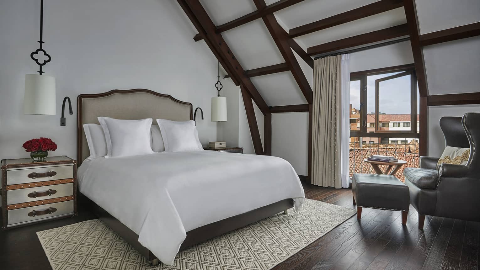 Hotel room with sloped beam ceilings, open balcony door, armchair, bed with white reading lamps, area rug