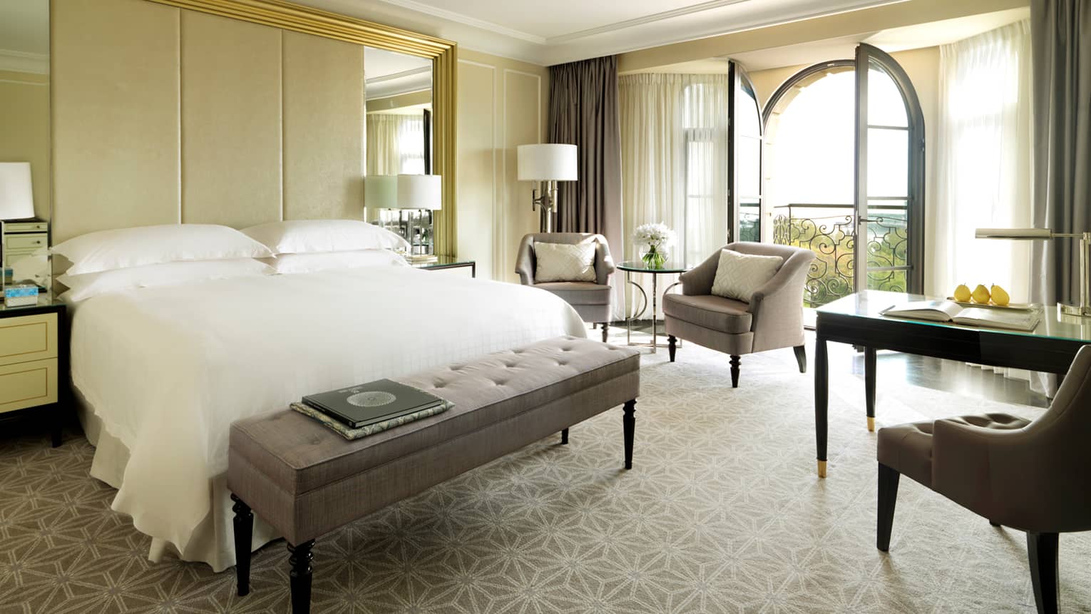 Sunny Deluxe hotel room with arched French doors, two armchairs, desk, bed with bench, books