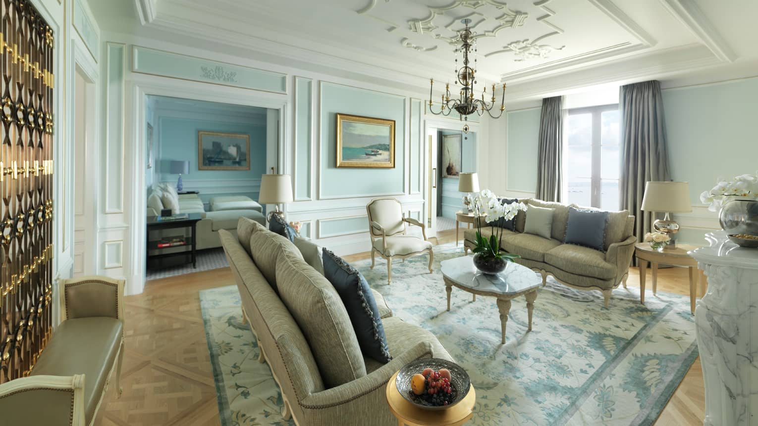 Presidential Suite sunny living room with two elegant sofas, coffee table under small chandelier by window, marble fireplace