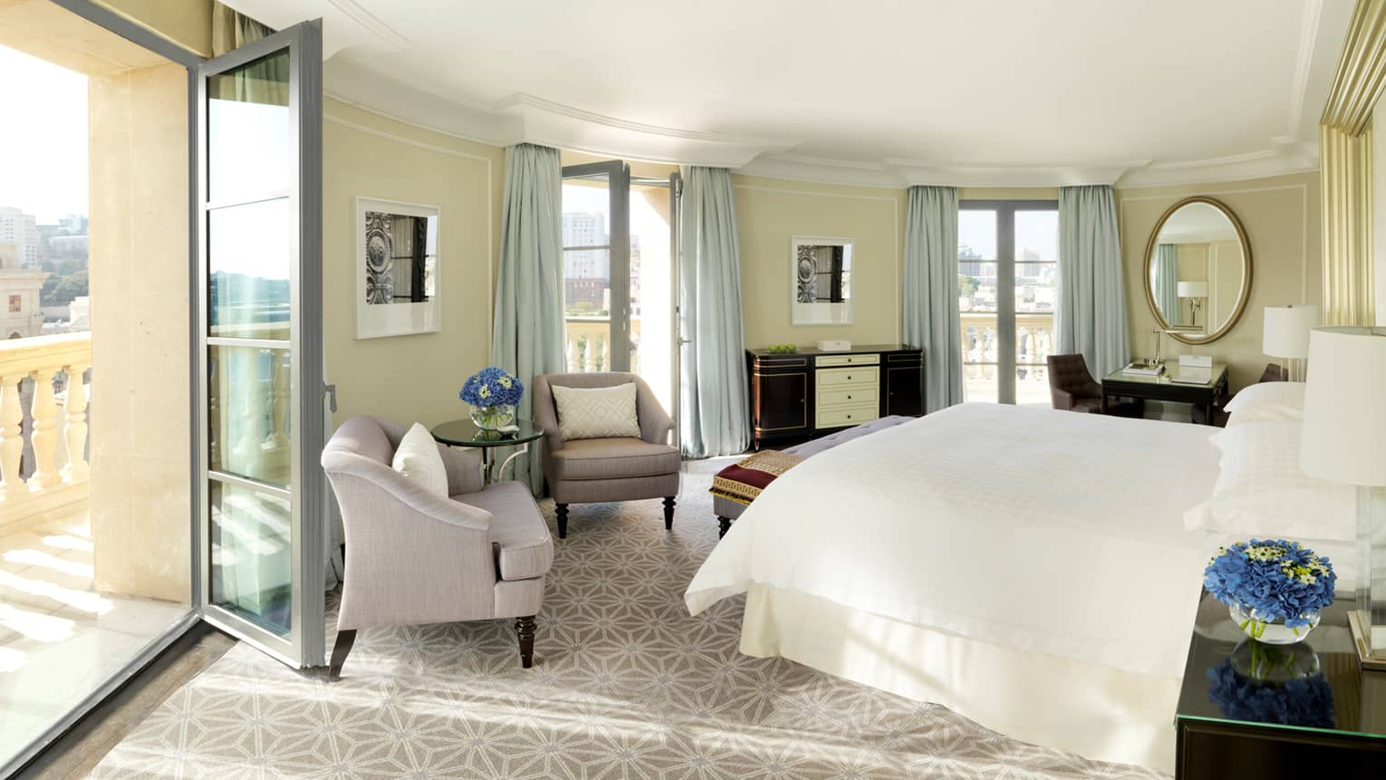 Sun streams in from open balcony door to curved hotel room with bed, plush armchairs, mirrors