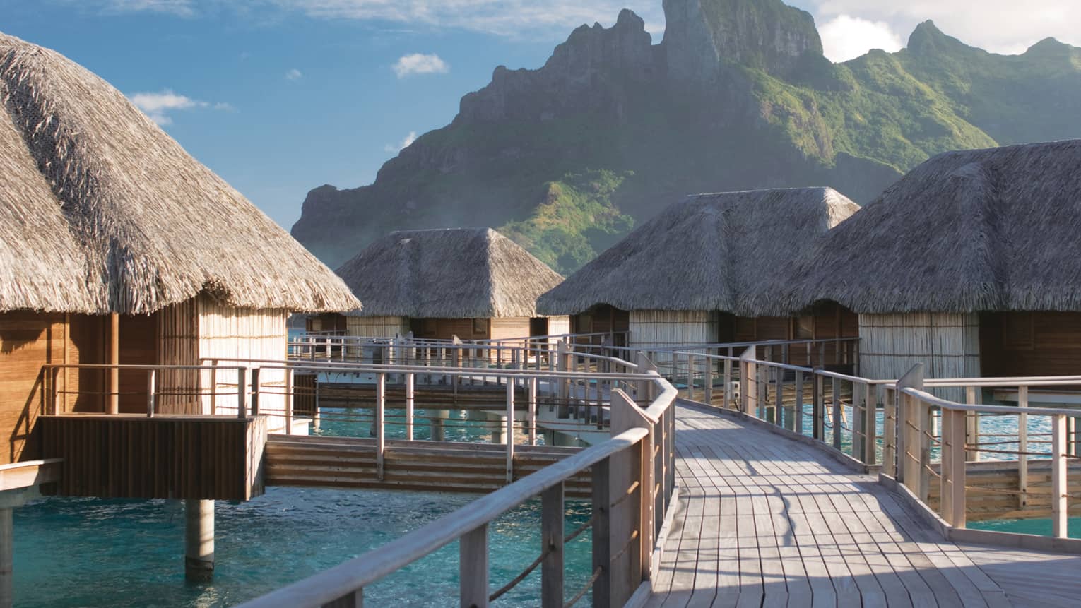 Curved deck connecting overwater bungalows with thatched roofs above lagoon
