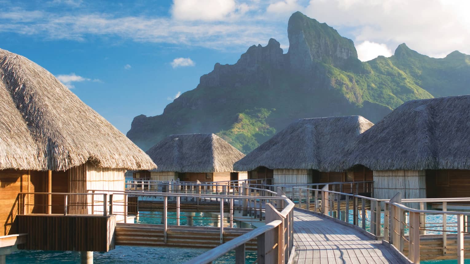 Curved deck connecting overwater bungalows with thatched roofs above lagoon