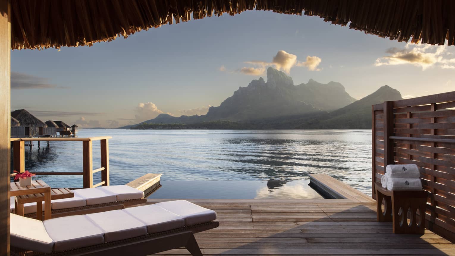 A view from a wooden Water Bungalow with white cushioned lounge chairs overlooking the ocean and mountains in the background at Four Seasons Bora Bora