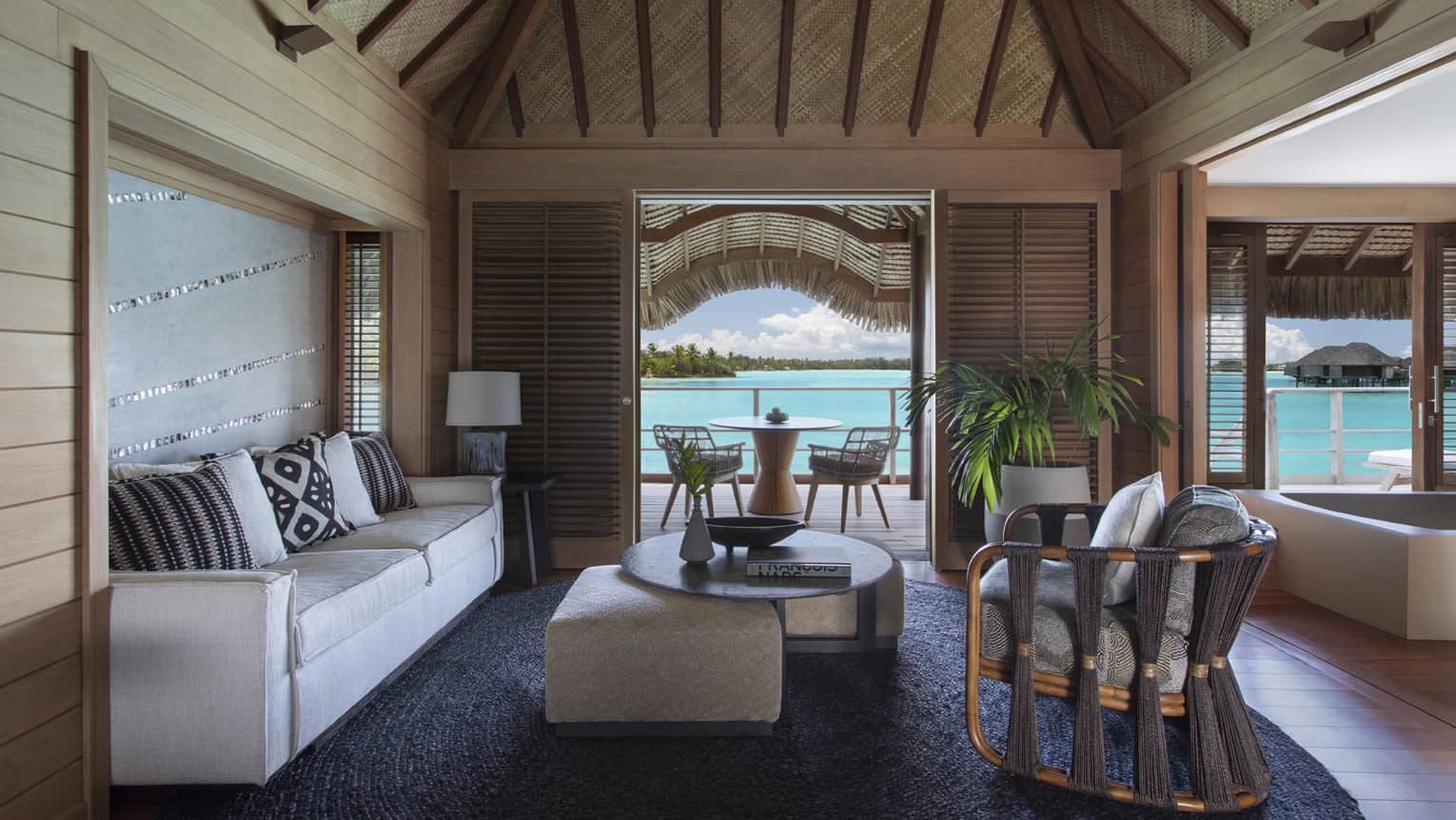 Overwater bungalow living area with neutral furniture, wood beams, tropical plants, and a patio with a view of the turquoise lagoon. 