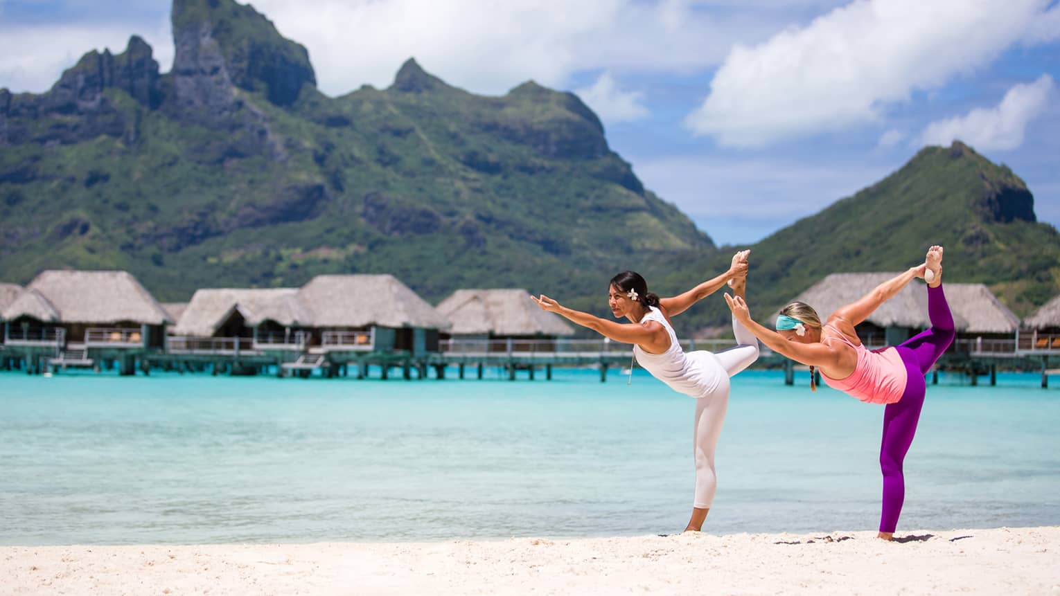 Two people practicing yoga on a tropical beach with overwater bungalows and mountains in the background