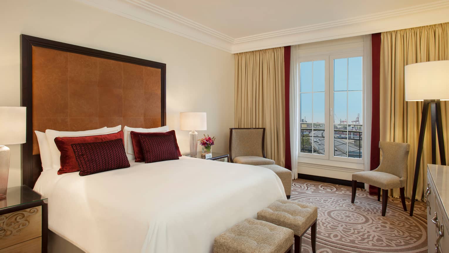 City-View One Bedroom Suite bed with tall padded leather headboard, small red accent pillows, two chairs by window
